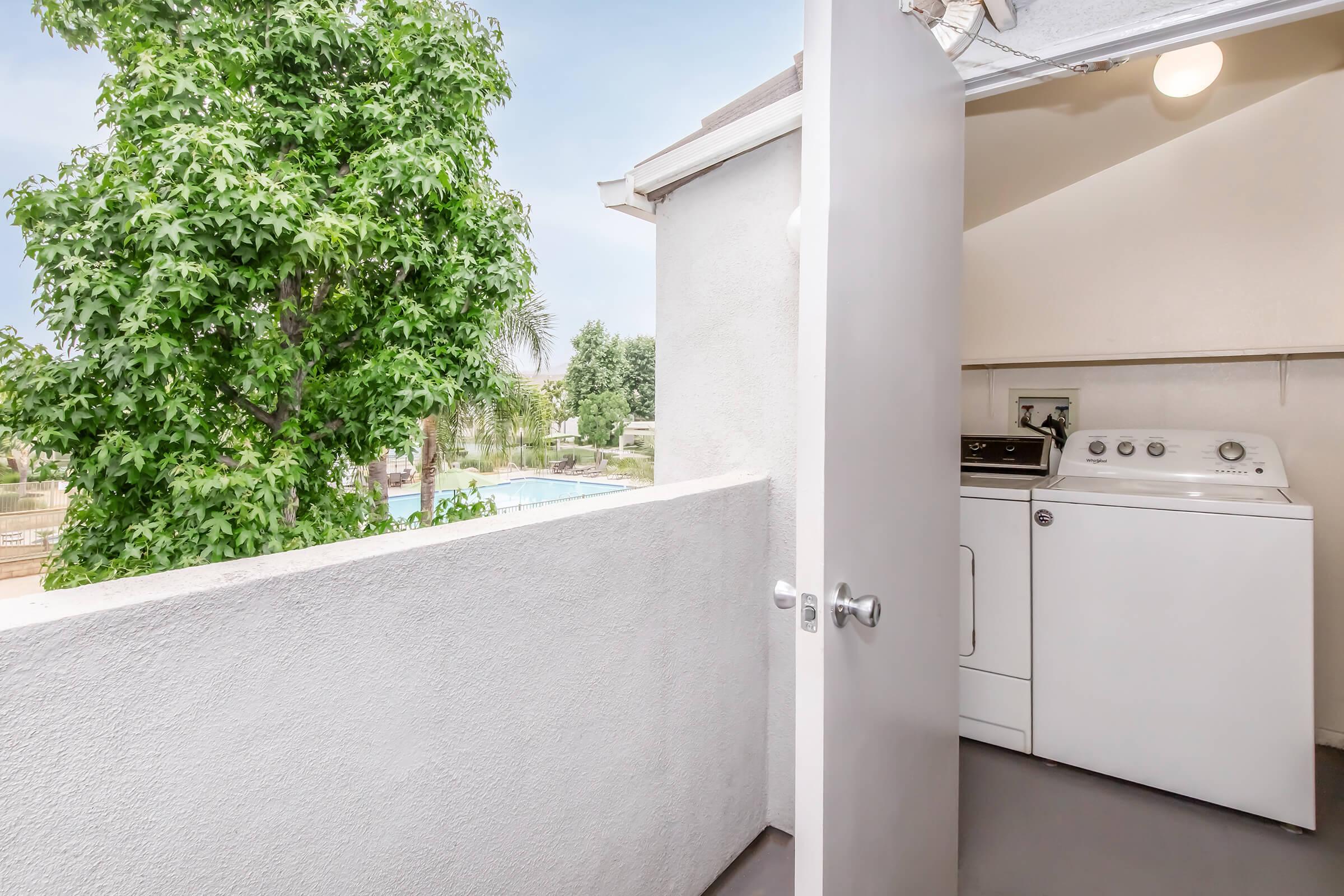 A laundry area with a washing machine and dryer, located on a patio or balcony. The door is open, revealing a view of a green tree and a pool in the background, suggesting a bright and sunny outdoor space.