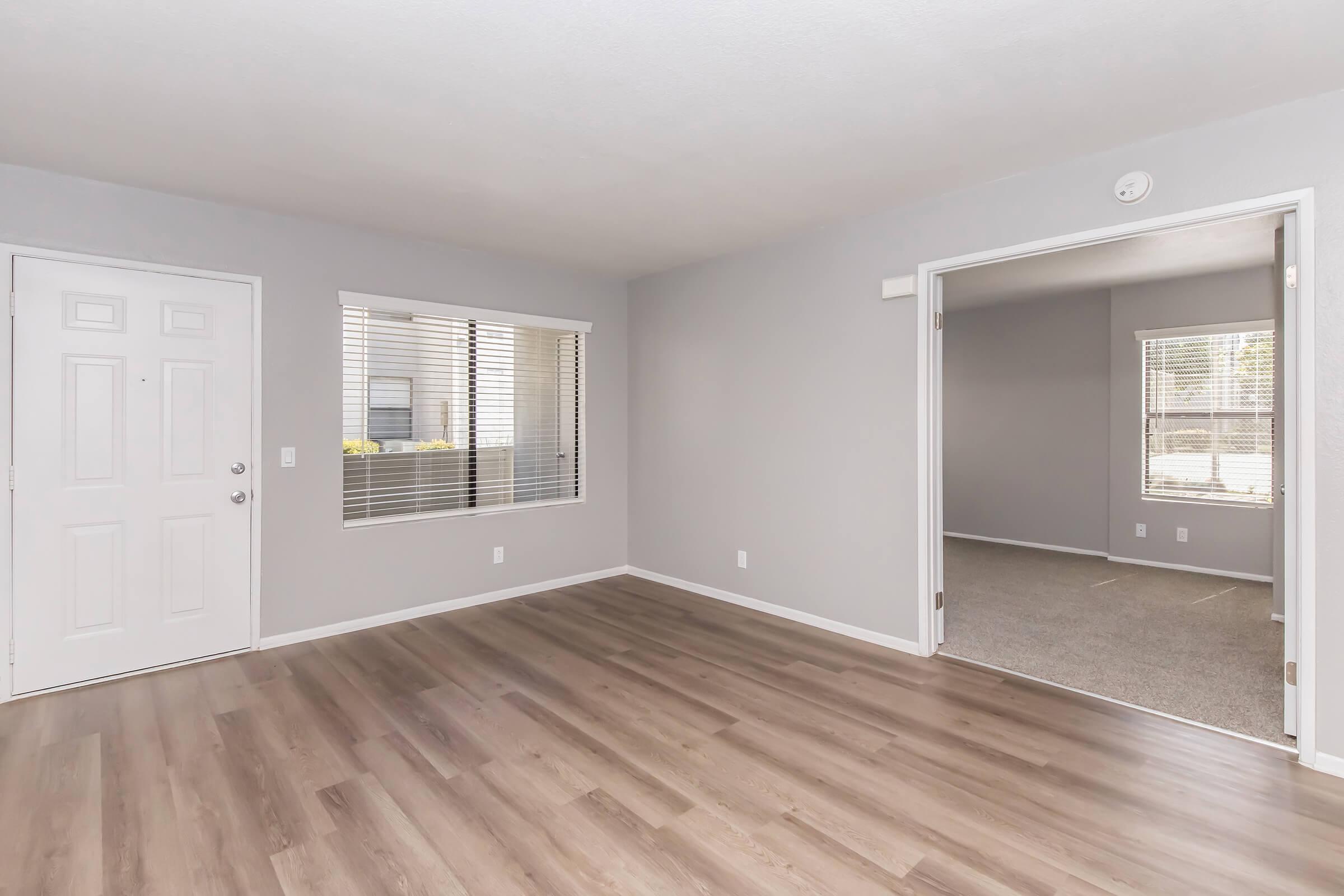 Spacious interior of a modern living room featuring light gray walls, wood laminate flooring, and large windows with blinds. The view includes a front door and an adjoining room with carpet. Natural light brightens the space, creating a welcoming atmosphere.