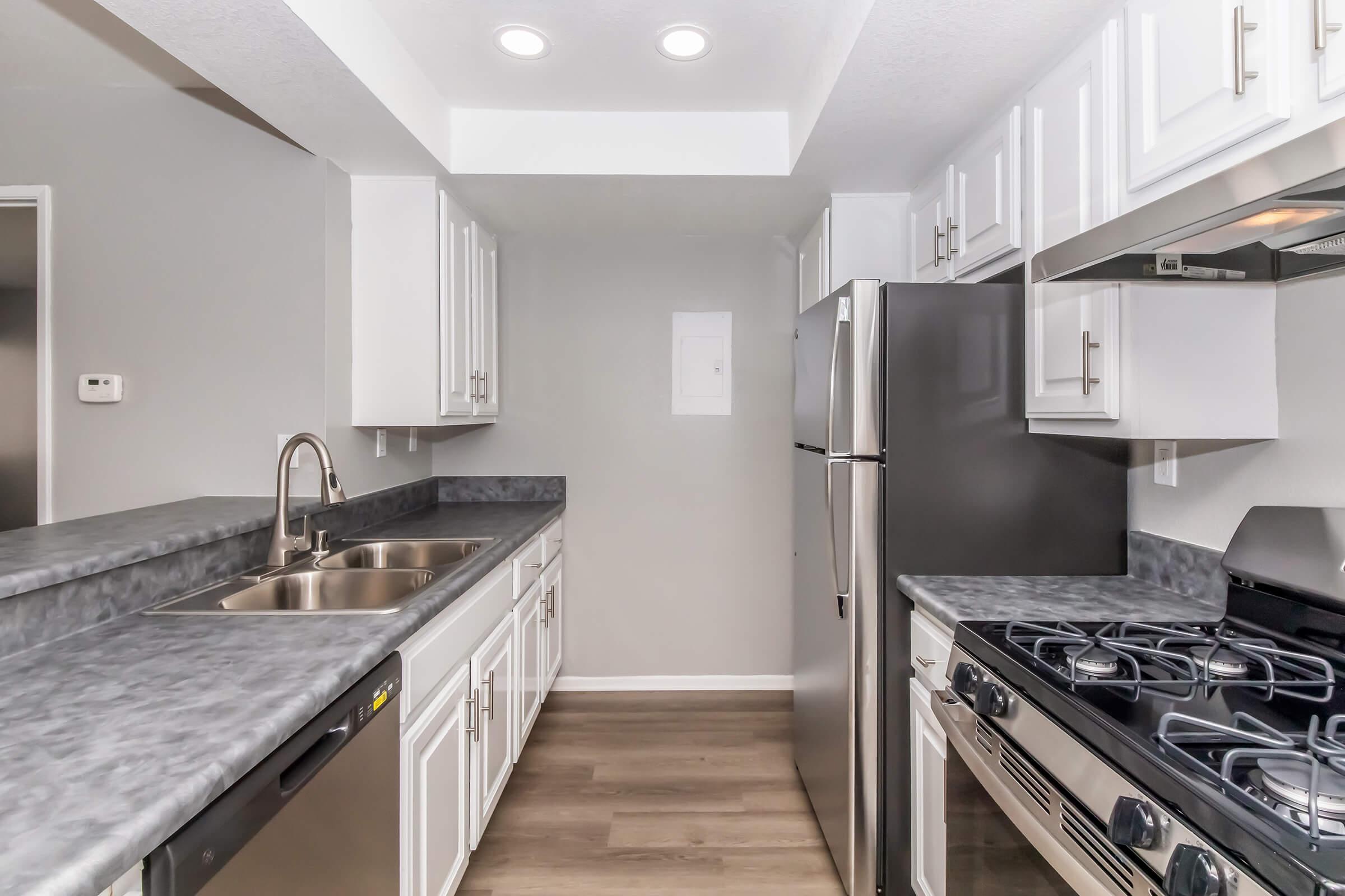 A modern kitchen featuring gray countertops, stainless steel appliances including a refrigerator and gas stove, white cabinetry, and pendant lighting. The space is well-lit, with an open layout and a sleek design, showcasing a clean and organized culinary environment.