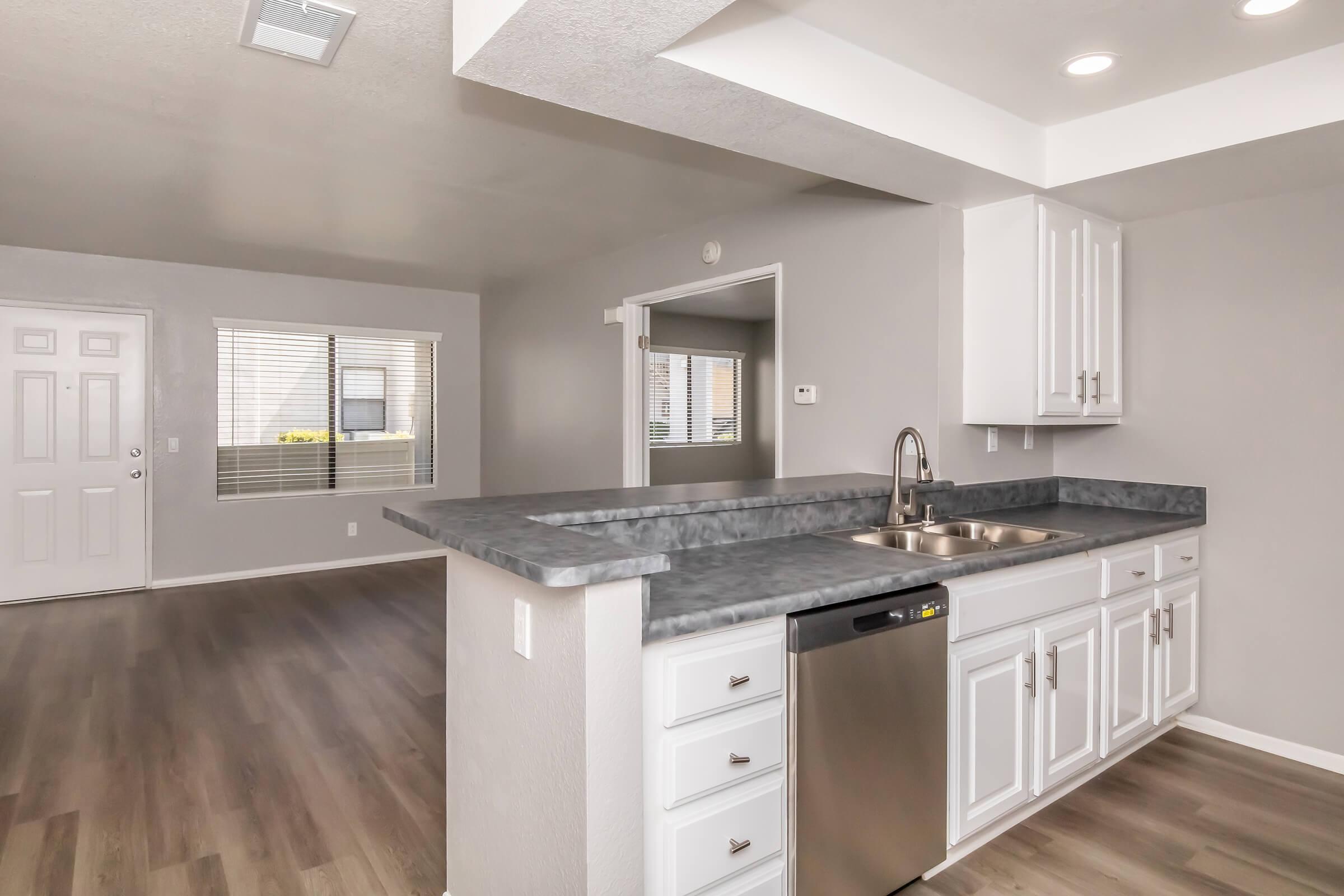 Modern kitchen with a gray countertop and stainless steel dishwasher. White cabinetry and overhead lighting create a bright atmosphere. The space opens to a living area with large windows, allowing natural light to fill the room. Hardwood flooring extends throughout, complementing the contemporary design.