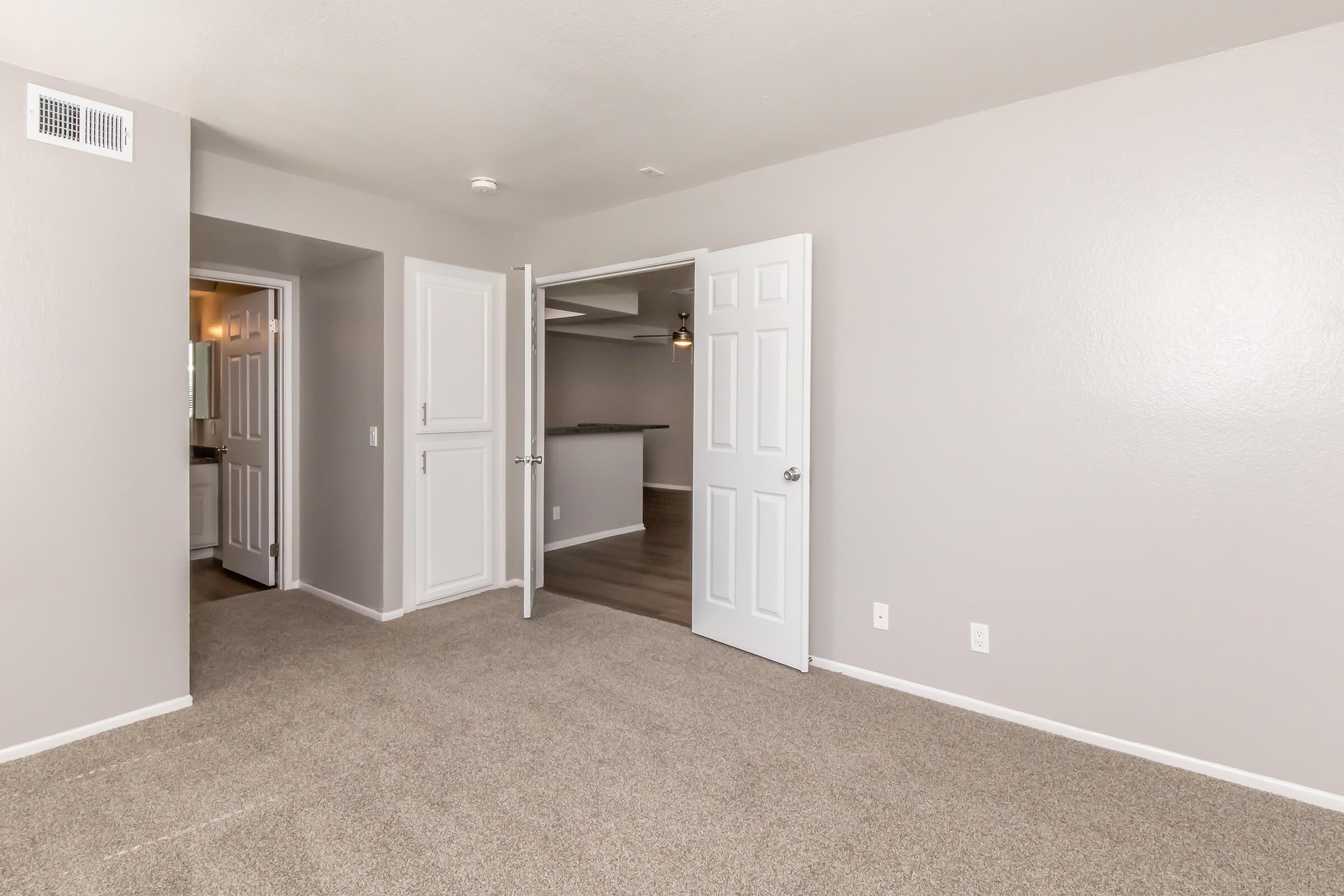 Empty room with light gray walls and carpeted flooring. Two white doors are open, leading to adjacent spaces. The left door opens to a bathroom, while the right one leads to another area. The overall atmosphere is bright and neutral, suitable for various interior designs.