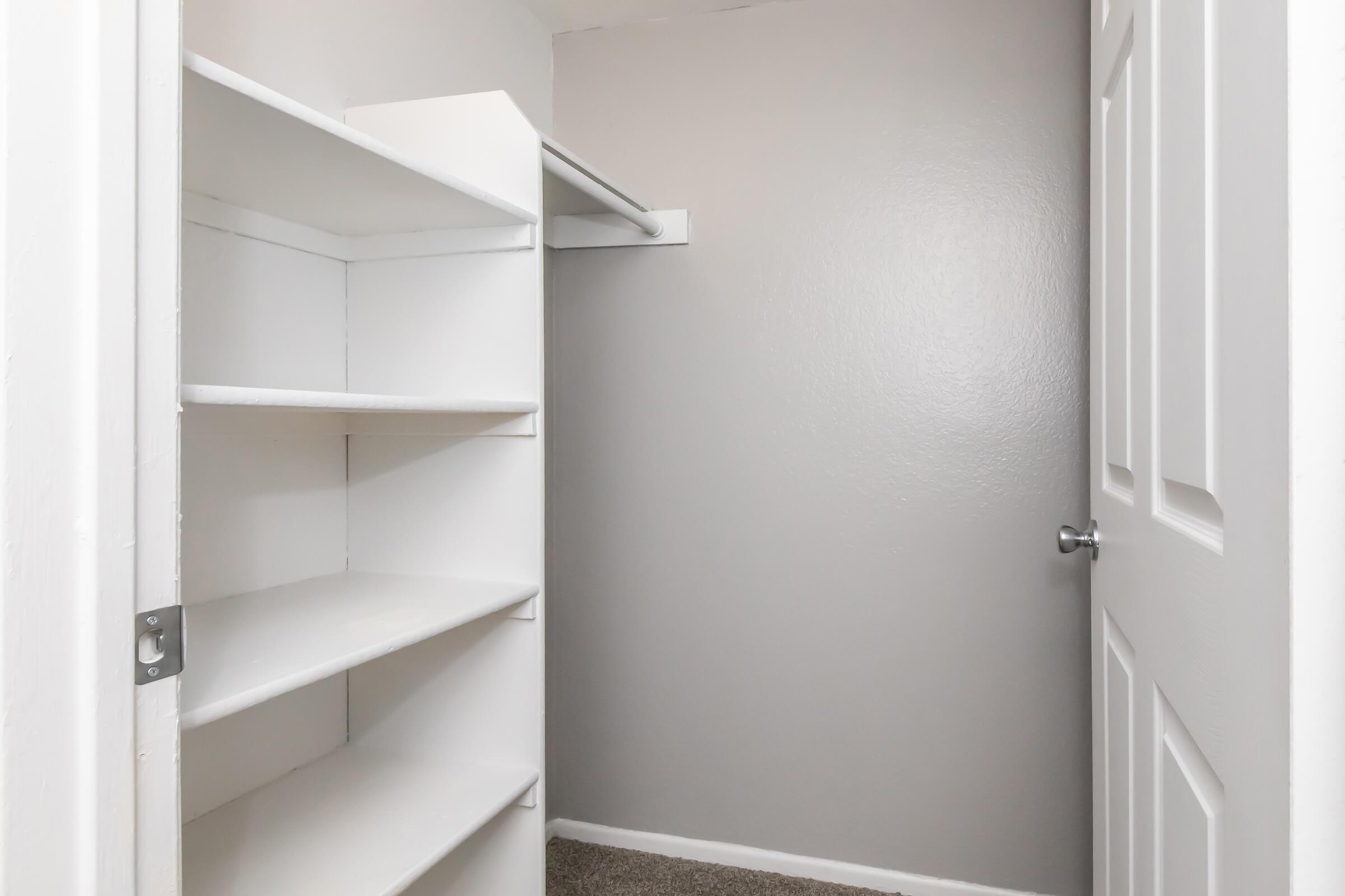 Empty closet with a gray wall, featuring white shelves on the left side and a hanging rod above for clothes. The door is partially open, revealing a clean, organized space with carpeted flooring.
