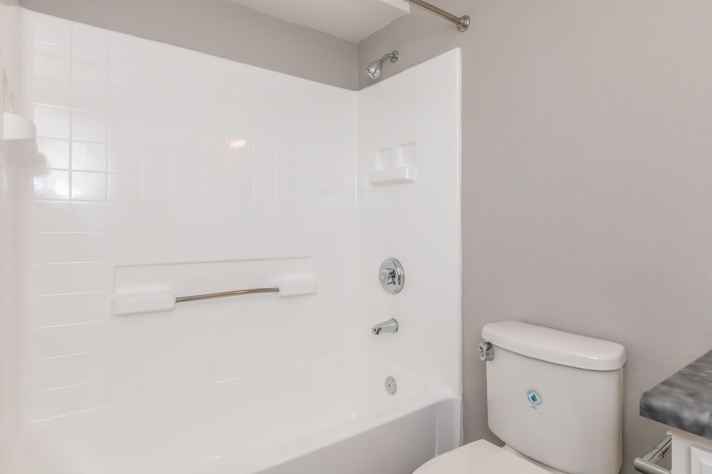 A clean bathroom featuring a white tiled bathtub with a shower, a grab bar, and a modern toilet. The walls are painted in a light gray color, providing a simple and contemporary aesthetic. The overall design emphasizes a functional and uncluttered space.
