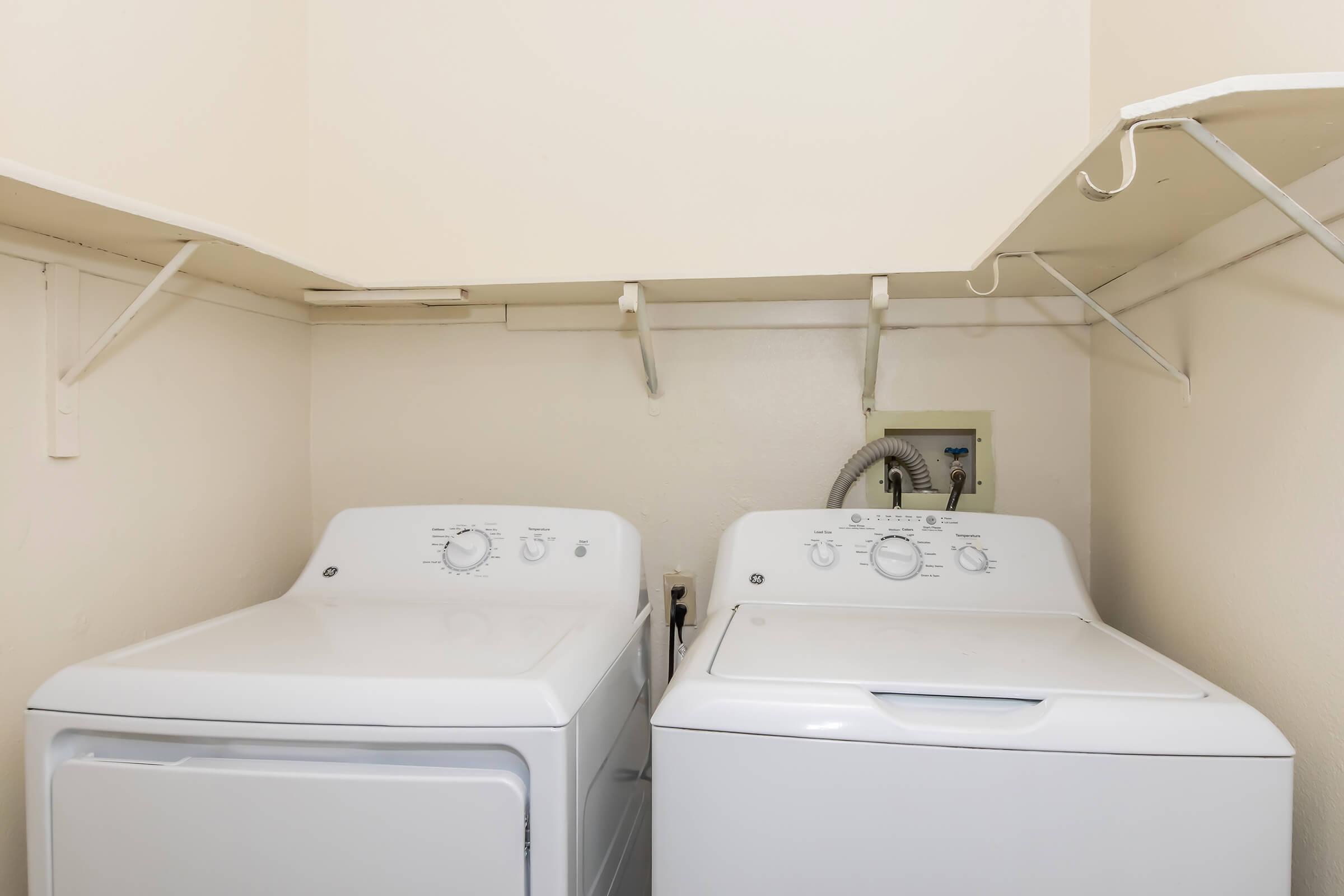 A laundry room featuring a white washing machine and a white dryer side by side. Above the appliances, there are shelves mounted on the wall for storage. The walls are painted a light color, creating a clean and simple environment.