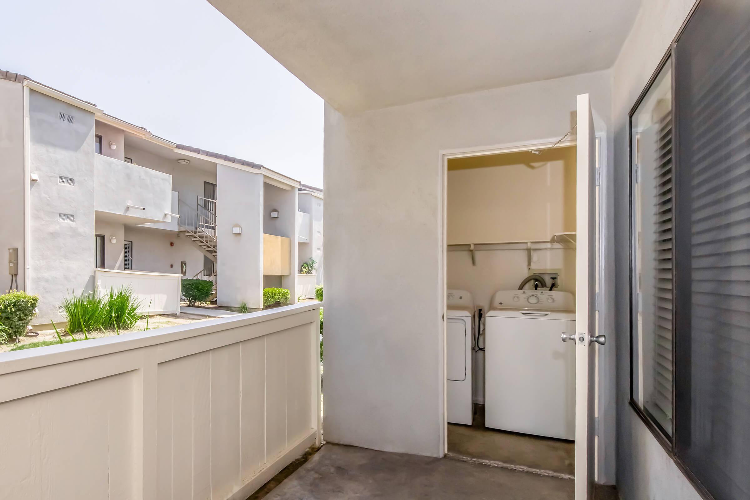 View from a balcony overlooking a utility area with a door leading to a laundry room containing a washing machine and dryer. Nearby, there are two residential buildings visible, surrounded by grass and shrubs under a clear blue sky.