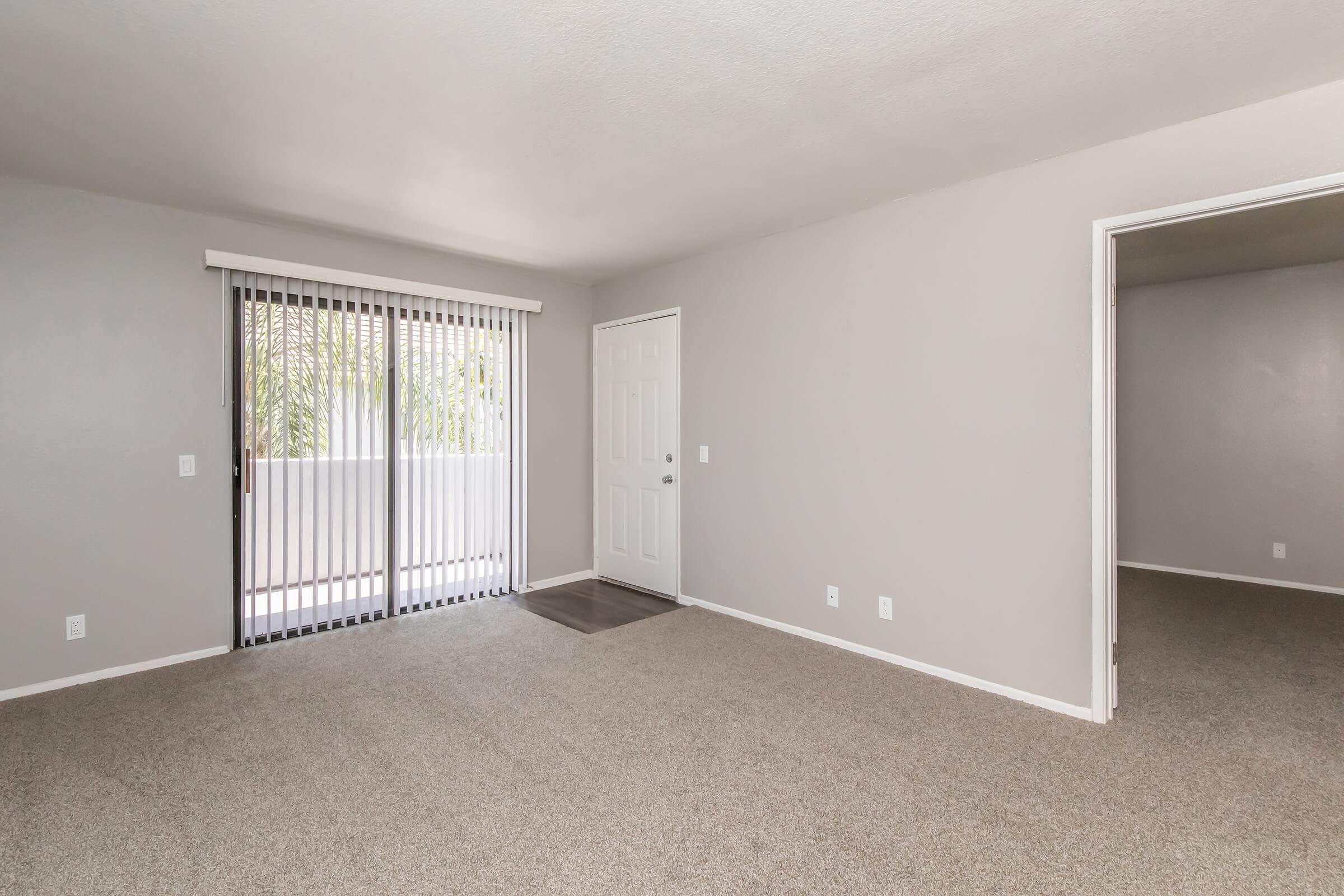 A spacious, empty room featuring light gray walls and carpet flooring. There are sliding glass doors with blinds leading to a balcony, and a door on the right side leading to another room. The overall ambiance is bright and minimalistic, suggesting potential for personalized decor.