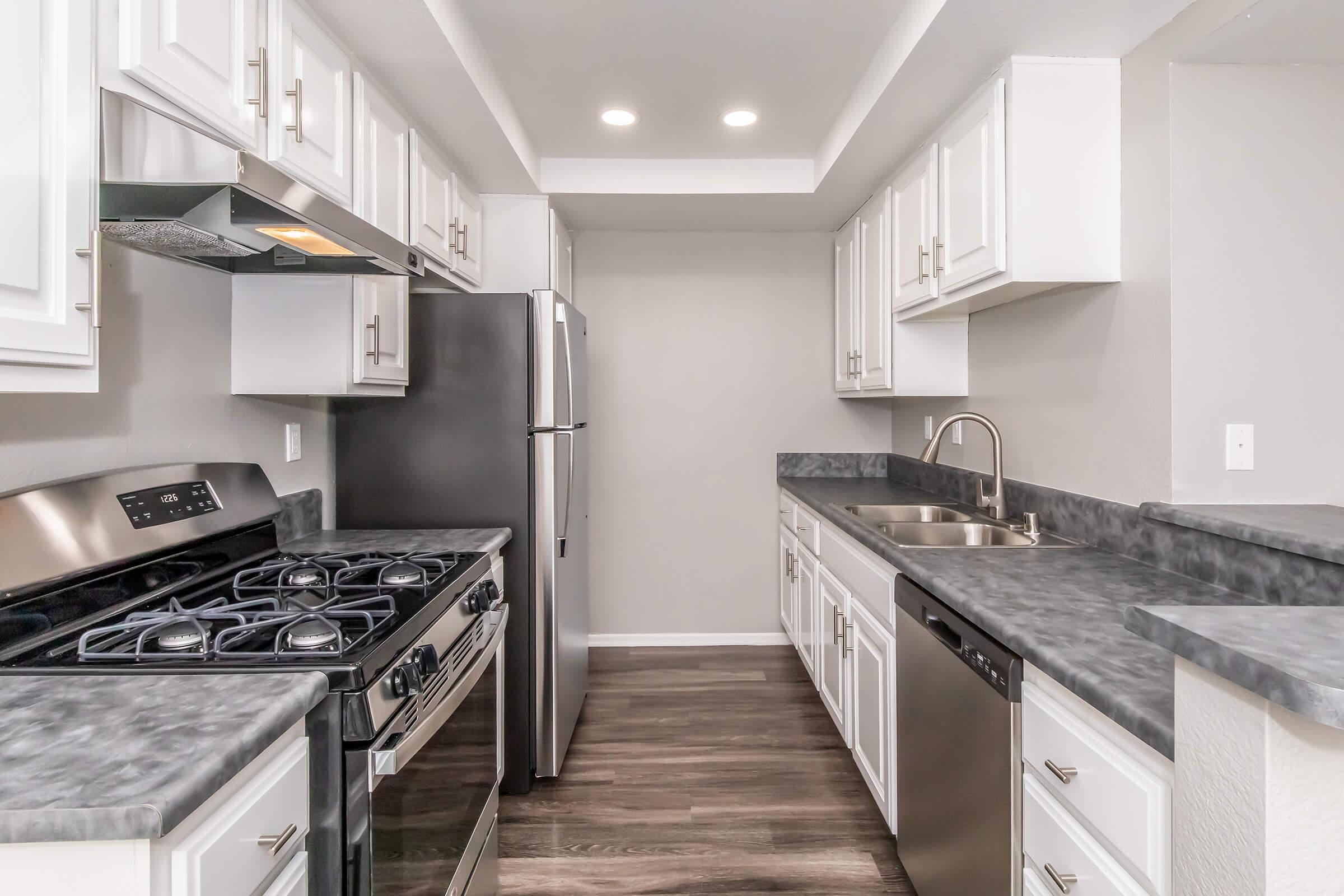 Modern kitchen featuring white cabinets, gray countertops, and stainless steel appliances. The space includes a gas stove, refrigerator, sink, and dishwasher, all arranged in a functional layout with overhead lighting. The flooring is a dark wood finish, adding warmth to the contemporary design.