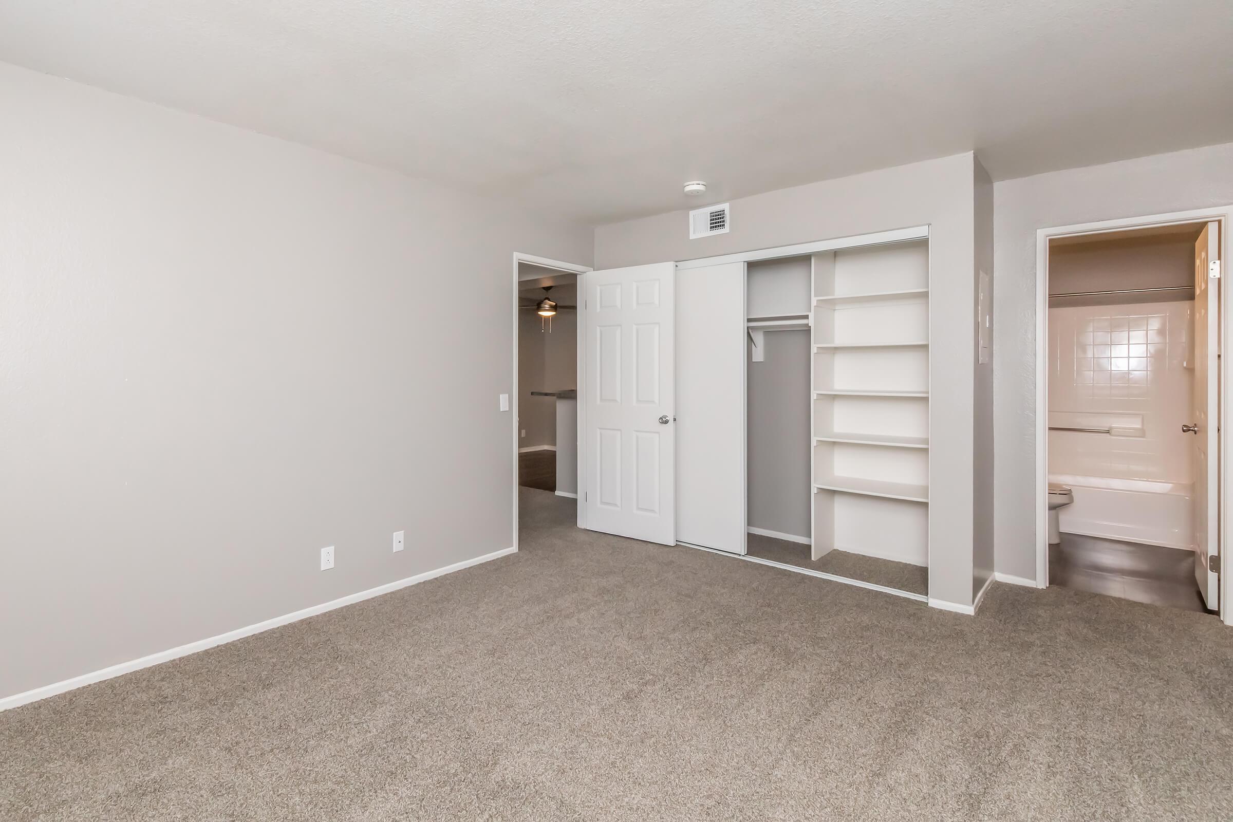 A spacious, empty room with light gray walls and beige carpet. It features a closed closet with sliding mirrored doors on the left and doorways leading to a bathroom and another room. Soft overhead lighting creates a bright, inviting atmosphere.