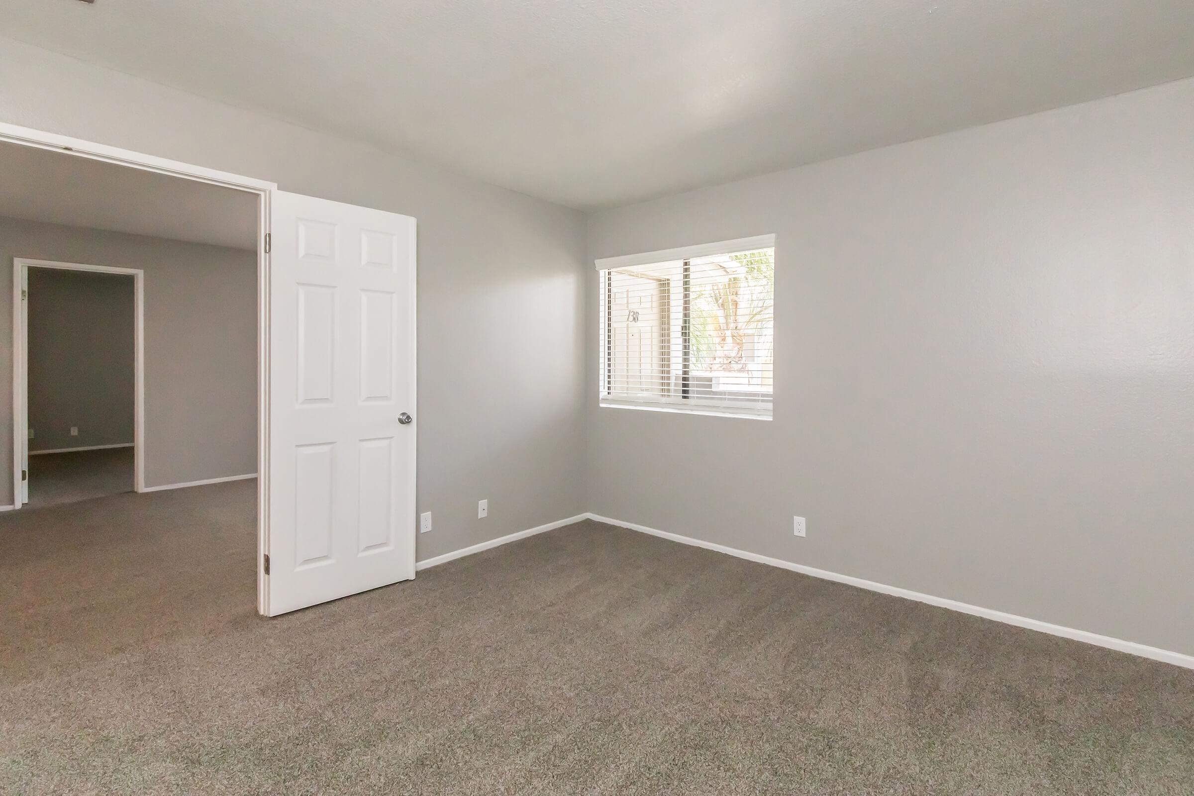 Empty room with light gray walls and carpet, featuring a window with blinds and a door partially open to another room. The space is well-lit, creating an airy atmosphere.