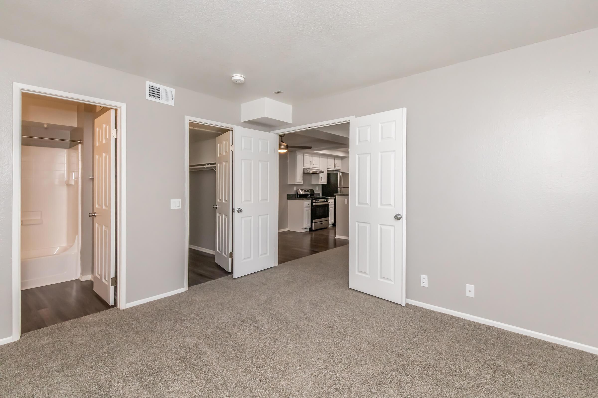 Spacious interior view of an apartment featuring two open doors leading to rooms, light gray walls, and a carpeted floor. A well-equipped kitchen is visible in the background, with modern appliances and an inviting layout. The image conveys a bright and airy living space.