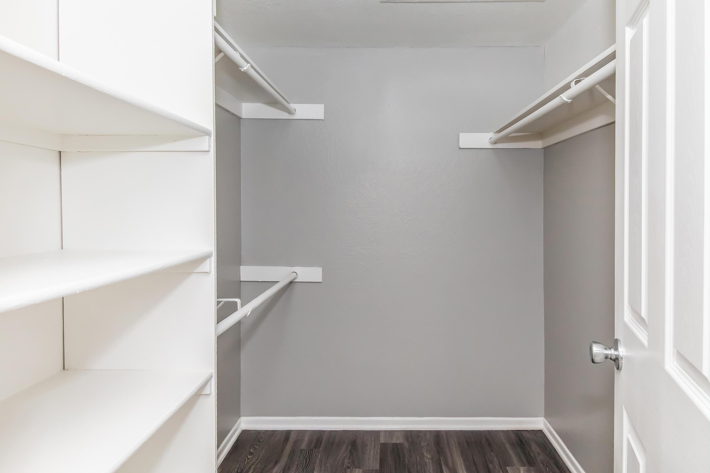 A vacant walk-in closet with gray walls, wooden flooring, and white shelving units. The closet features two hanging rods for clothing. The door is slightly ajar, allowing for a view of the empty space.
