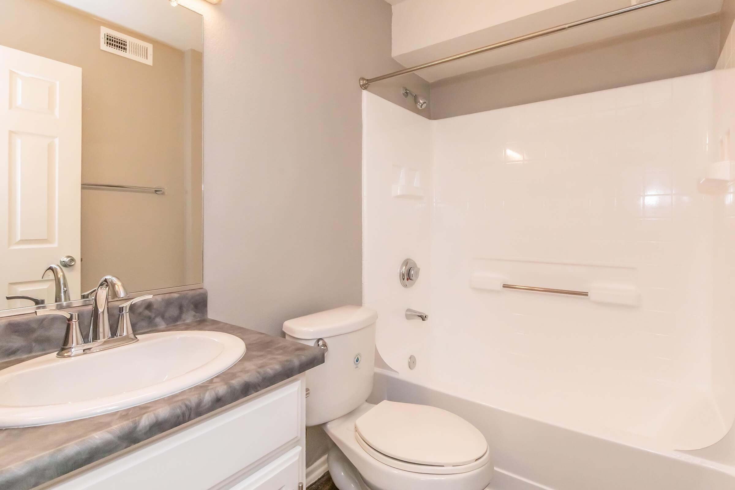 A clean and modern bathroom featuring a white bathtub with a shower, a toilet, and a sink with a marble countertop. Light gray walls and neutral-colored fixtures create a bright and inviting atmosphere.