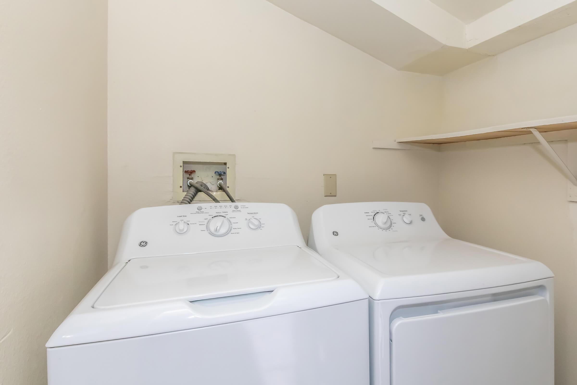 A laundry room featuring a white washing machine and a matching dryer side by side, with a wall-mounted hook-up panel in the background. A shelf is visible above the appliances, all set against light-colored walls. The space appears tidy and functional, ideal for laundry tasks.