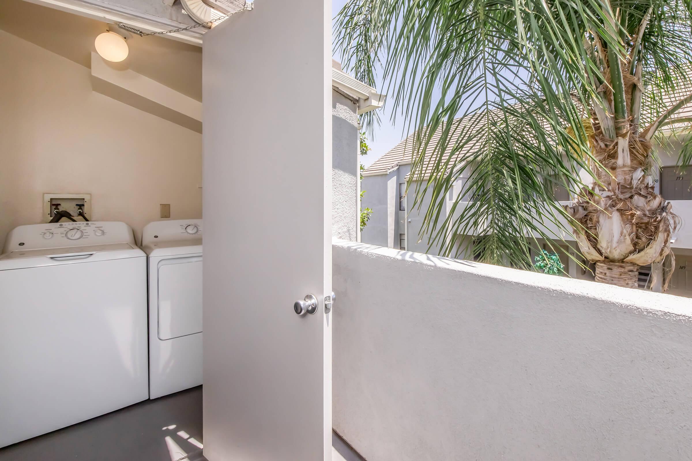 A doorway open to a balcony with a view of a palm tree. On the left, there are stacked white washing machines. The wall is light-colored, and the scene is bright with natural light coming in. The balcony overlooks a residential area with tiled roofs.