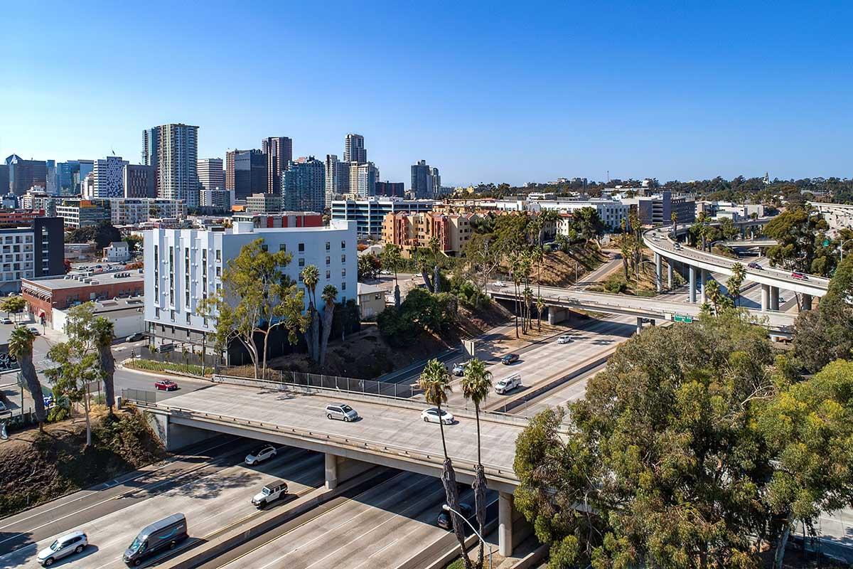 A panoramic view of a bustling urban landscape featuring several modern buildings and skyscrapers. In the foreground, multiple lanes of a freeway are visible with cars in motion.