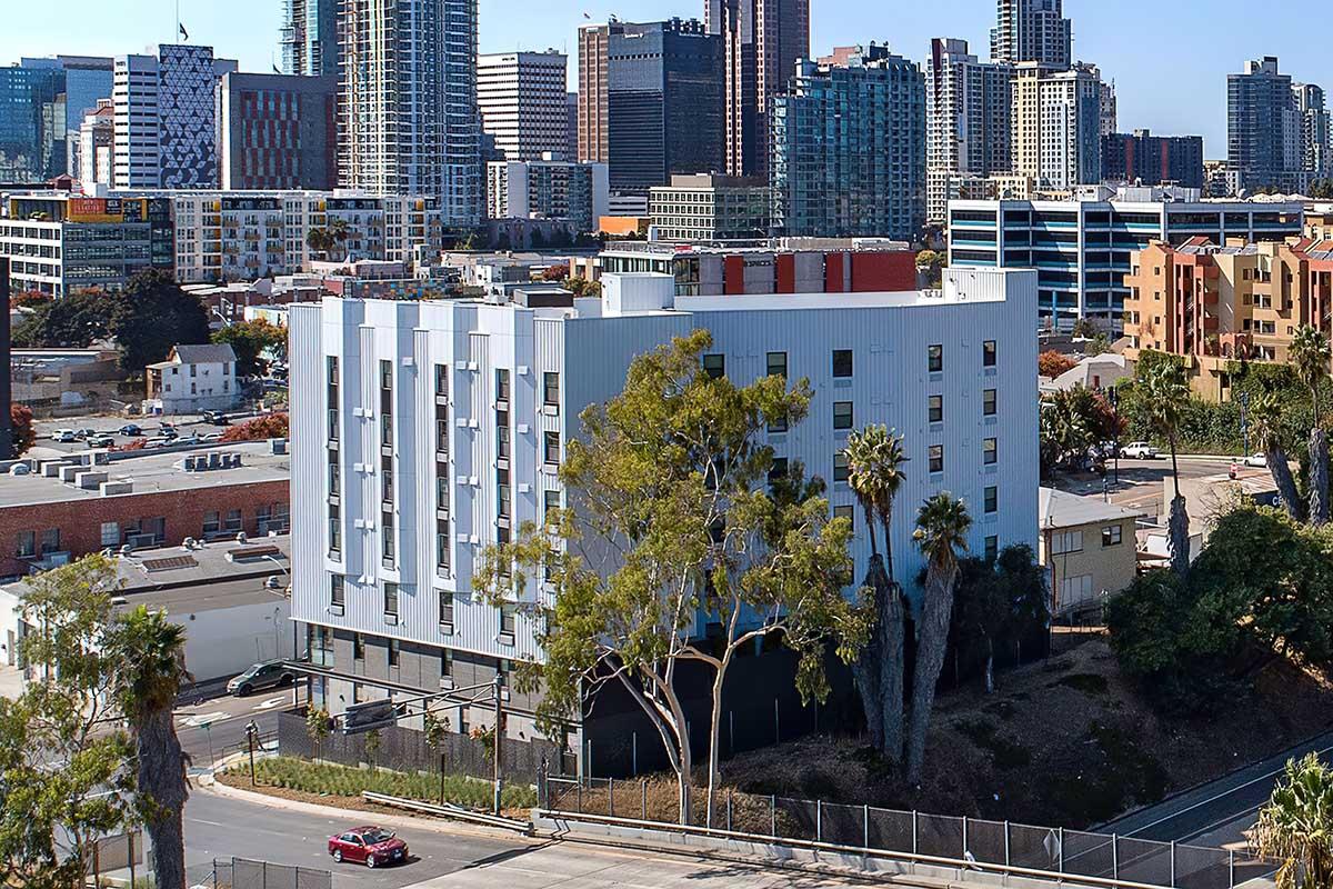 Cityscape view featuring a modern multi-story building surrounded by palm trees, with a backdrop of urban skyscrapers and a clear blue sky.