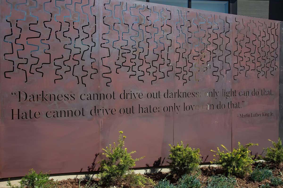 A metal wall featuring a quote by Martin Luther King Jr.: "Darkness cannot drive out darkness; only light can do that. Hate cannot drive out hate; only love can do that." The design includes a pattern of abstract lines and is surrounded by low vegetation.
