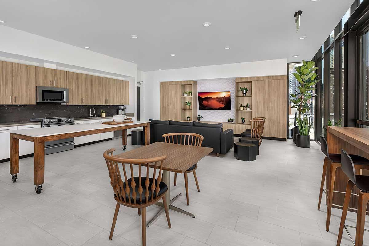 Modern open-concept lounge area featuring a sleek kitchen with wooden cabinetry and appliances, a cozy seating arrangement with a black couch, and wooden tables and chairs.
