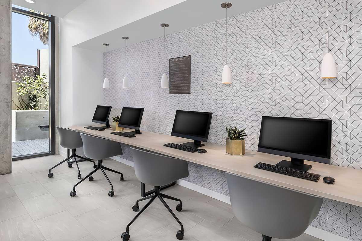 A modern workspace featuring four black desktop computers on a light wooden table. Each workstation has a gray rolling chair and a small plant.