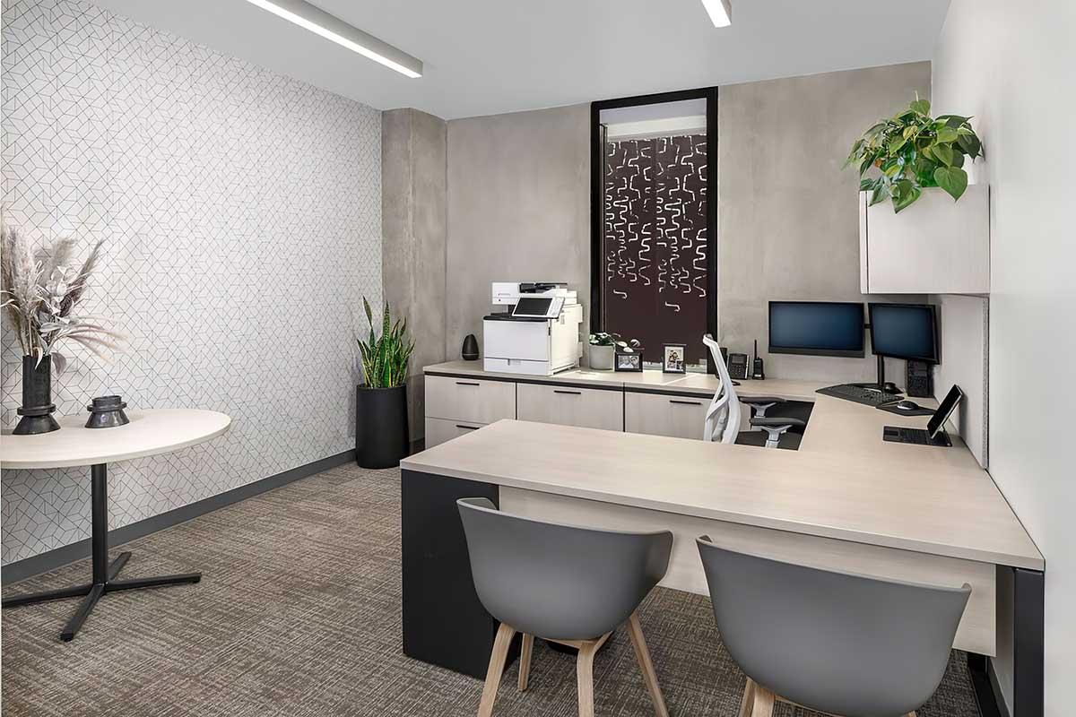 Modern office space featuring a desk with two computer monitors and a printer. A round table with chairs is visible, along with plants and decorative elements
