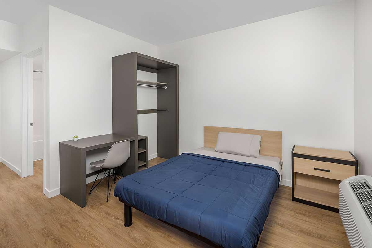 A simple, newly furnished bedroom featuring a bed with a blue comforter, a modern desk with a chair, a wardrobe, and a nightstand.