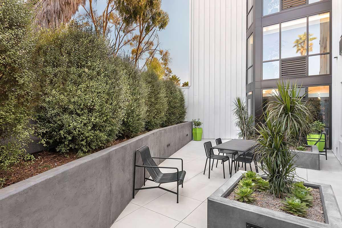 A modern outdoor patio area featuring a concrete wall, black metal chairs, a wooden table, and planters with greenery.