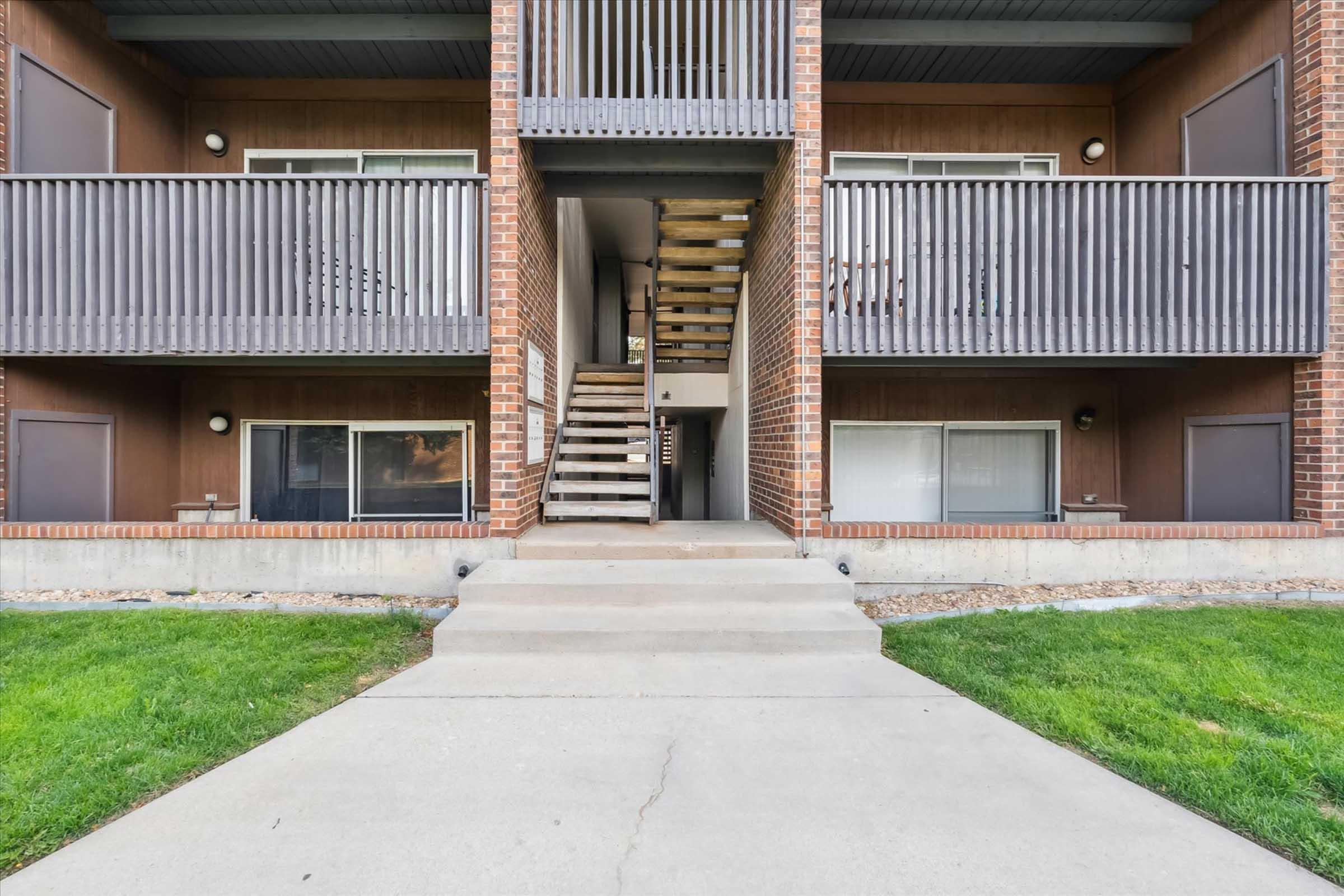 View of a multi-story residential building entrance with a staircase leading to upper floors. The ground level features two balconies with wooden railings, and a concrete pathway leads up to the entrance. The surrounding area has green grass and simple landscaping.