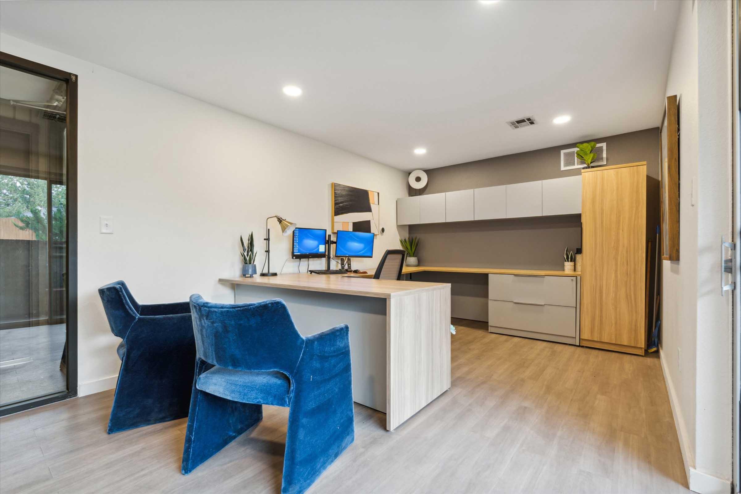 A modern home office featuring a wooden desk with dual monitors and a black chair. Two blue velvet chairs are positioned in front of the desk. The room has light-colored walls, ample lighting, and minimal decorations, creating a clean and professional workspace ambiance. A large window suggests natural light coming in.