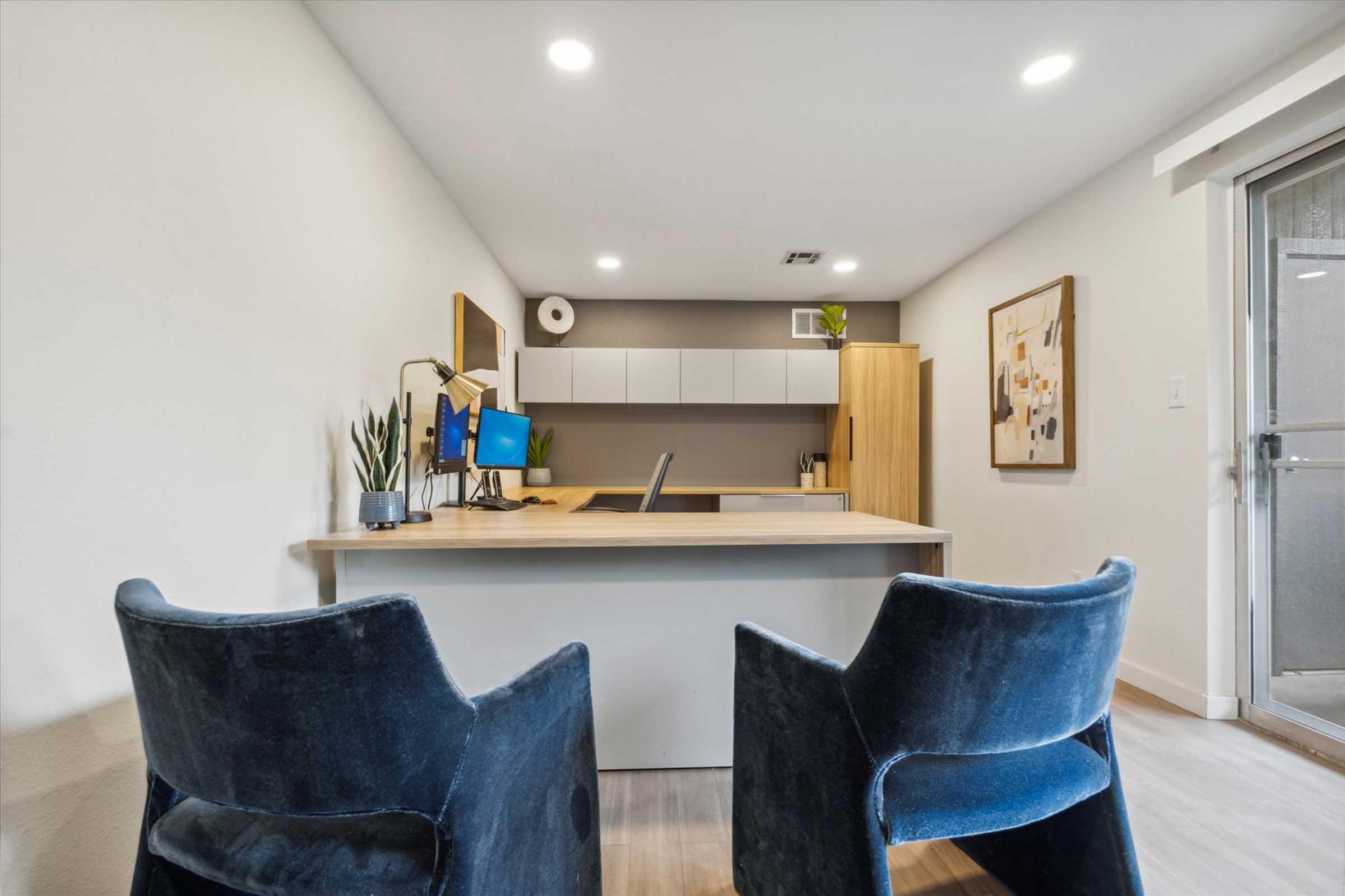 A modern office space featuring a wooden desk with a computer set up, two blue chairs, and light-colored walls. There are plants and decorative items on the desk and wall, creating a clean and organized atmosphere. A large window provides natural light.
