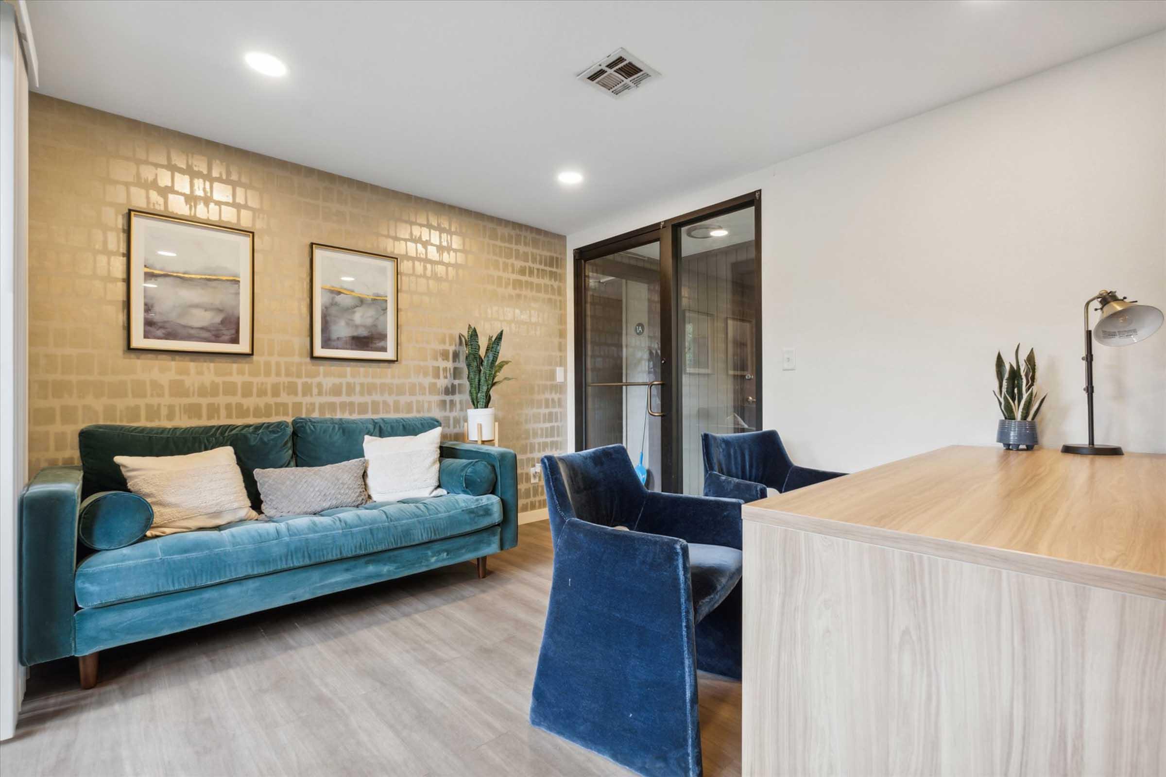 A modern waiting area featuring a teal velvet couch with cushions, two blue upholstered armchairs, and a wooden reception desk. The walls are decorated with framed art, and a potted plant adds a touch of greenery. Large glass doors lead to another space, enhancing the contemporary feel.