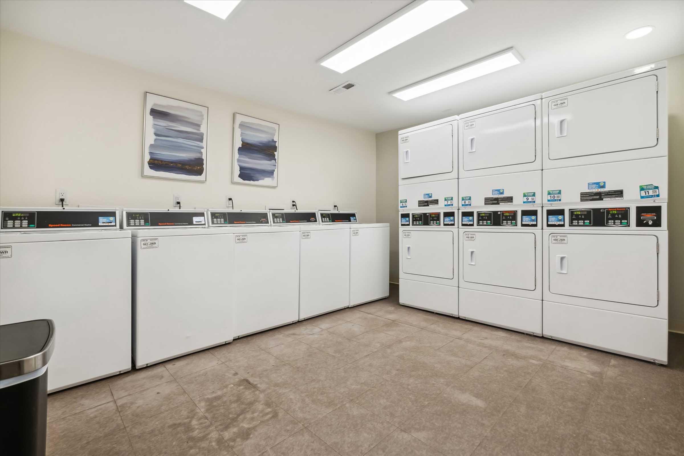 A modern laundry room featuring multiple white washing machines and dryers arranged in two rows. The machines have digital displays and are set against a neutral wall. Above them, two abstract paintings are hung. The floor is tiled and there is a trash can visible in the corner. Bright overhead lighting illuminates the space.