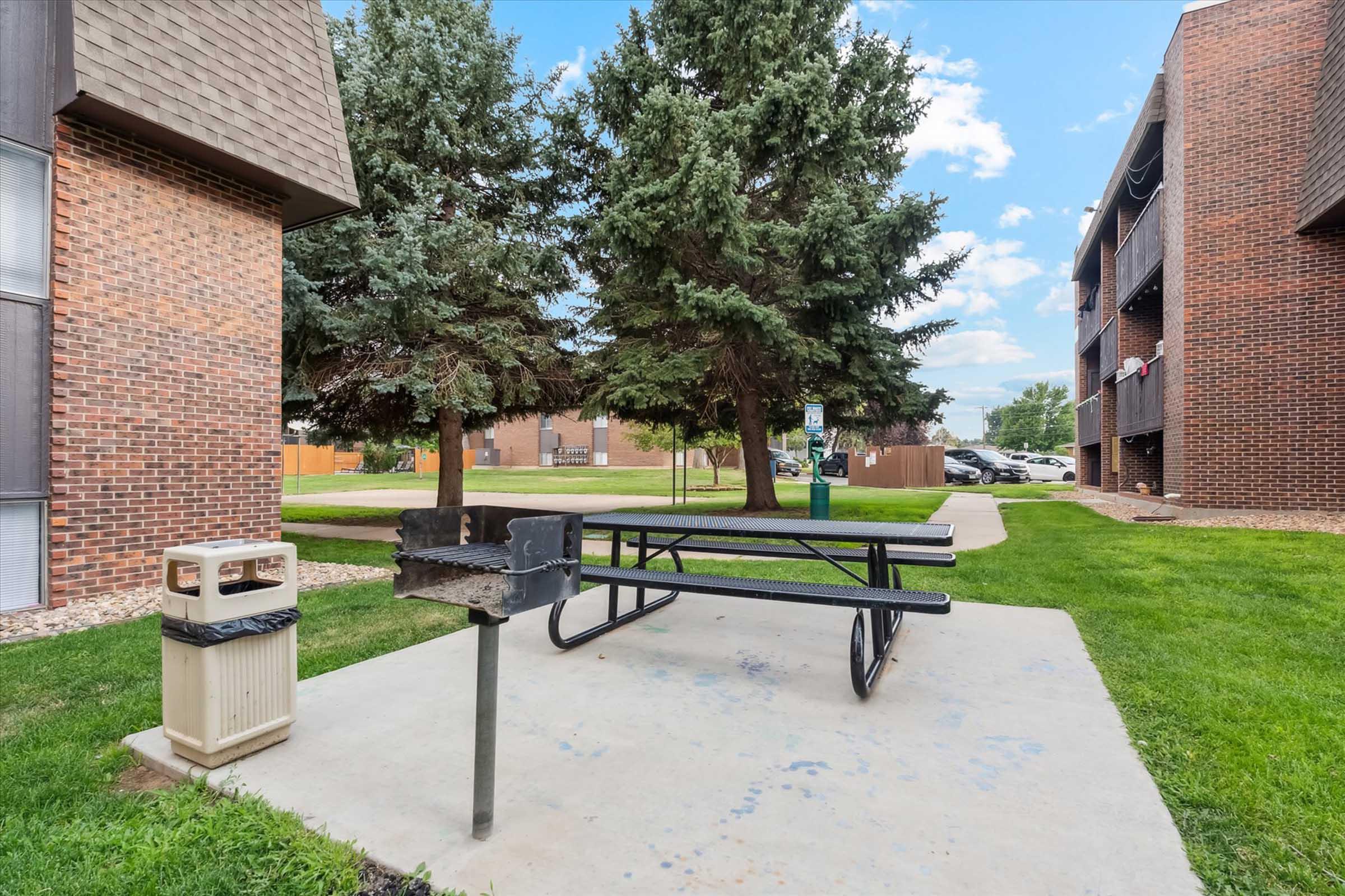 A picnic area featuring a metal grill and a large picnic table surrounded by grassy lawns and trees. Nearby, there is a trash bin and multiple apartment buildings in the background under a partly cloudy sky.
