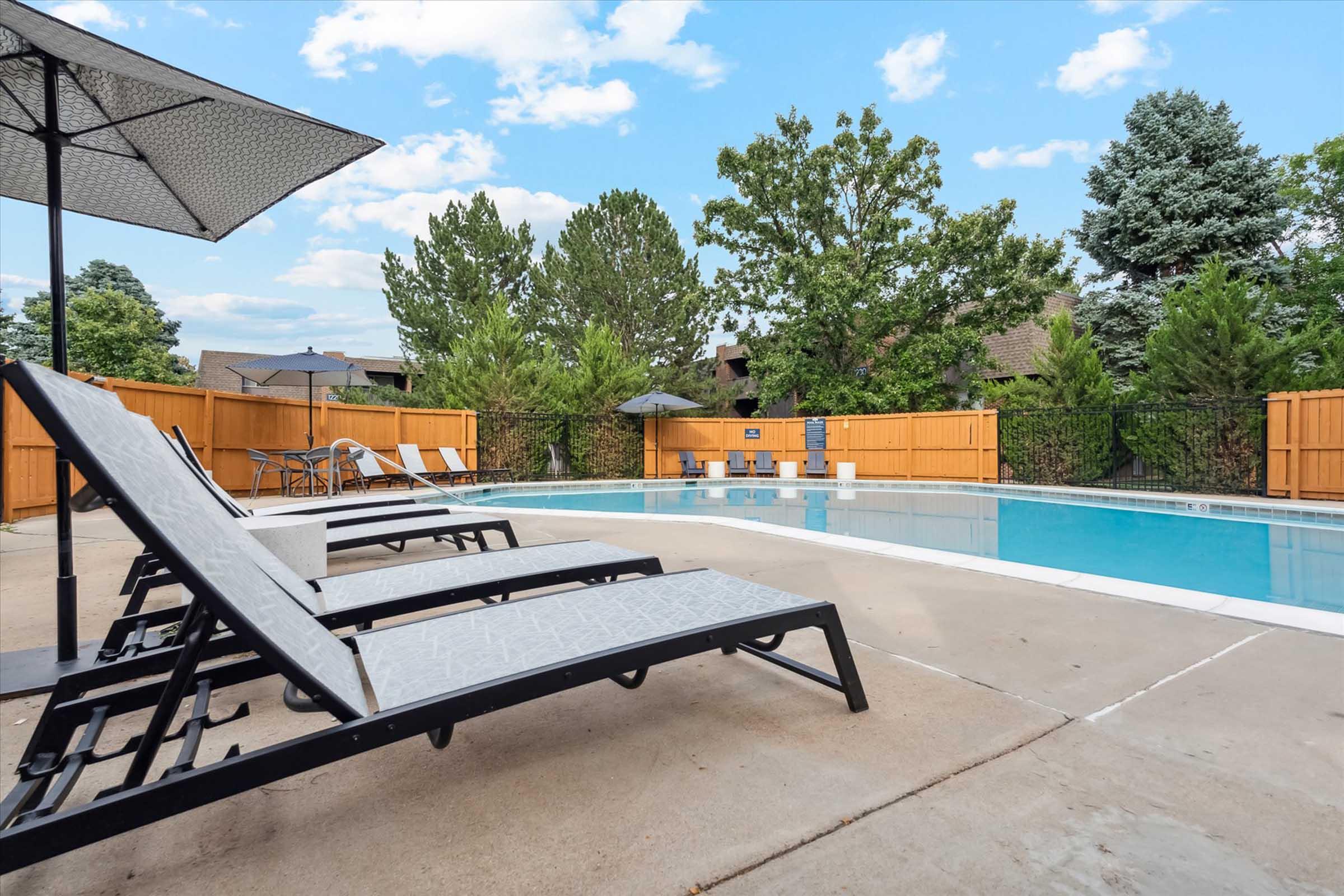 A swimming pool surrounded by lounge chairs, with umbrellas for shade. The area features a wooden fence and greenery in the background, under a bright blue sky with scattered clouds. Ideal for relaxation and outdoor enjoyment.