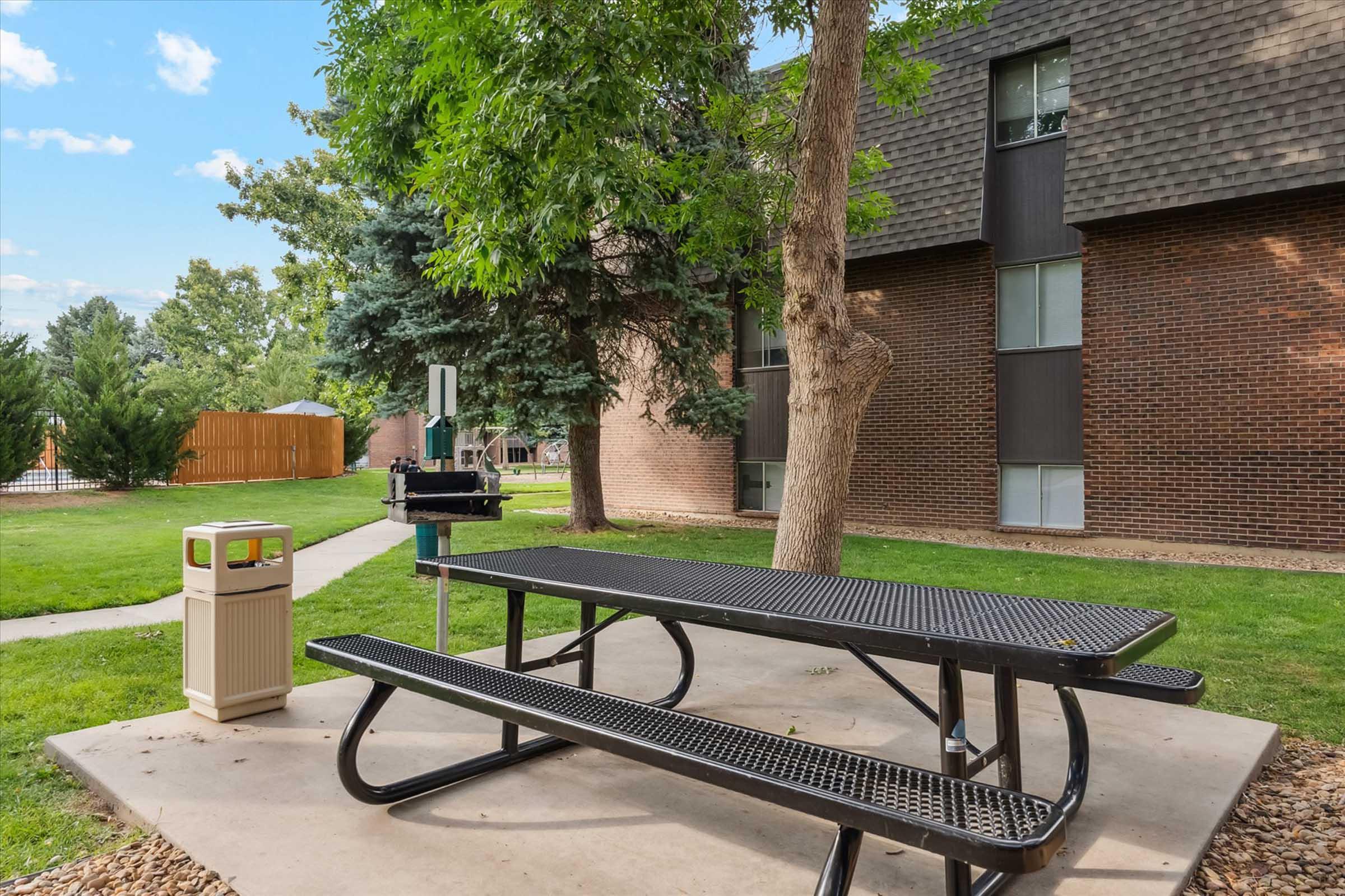 A picnic area featuring a black metal table with attached benches, a charcoal grill nearby, and a trash can. The setting includes grassy lawns and trees, with a brick building in the background. It’s a sunny day with a few clouds in the sky.