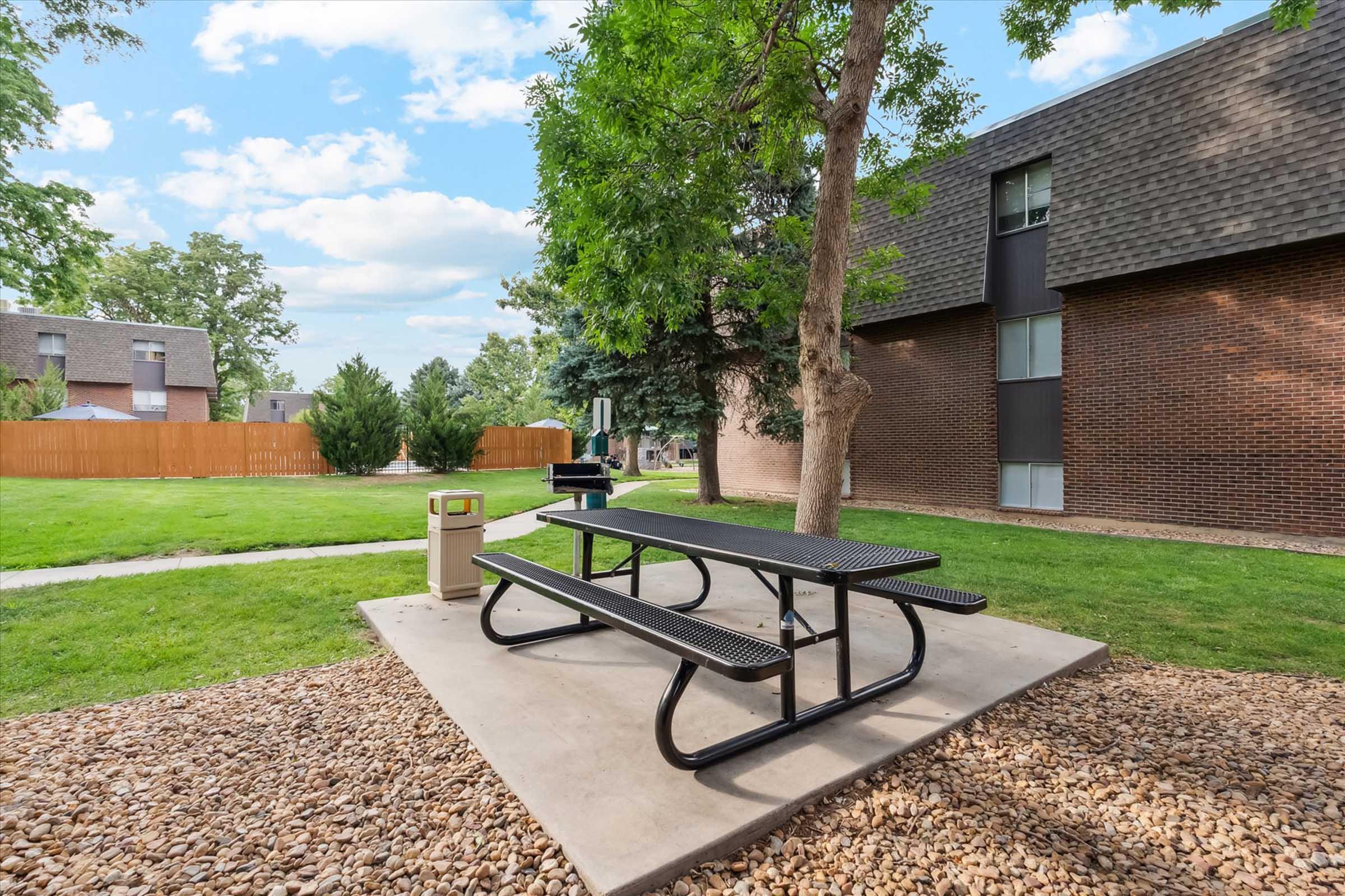 A picnic area featuring a black metal table with attached benches on a concrete slab, surrounded by a grassy lawn and rock landscaping. In the background, there is a tree and a brown brick building, along with a trash can and a grill, against a partly cloudy sky.
