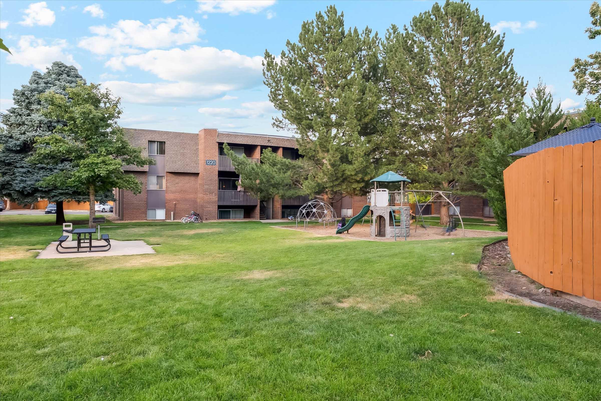 A grassy outdoor area featuring a playground with climbing structures, surrounded by trees and an apartment building. There is a seating area with a bench and a wooden fence visible in the background. The setting is bright and sunny, creating a welcoming atmosphere for residents and children.