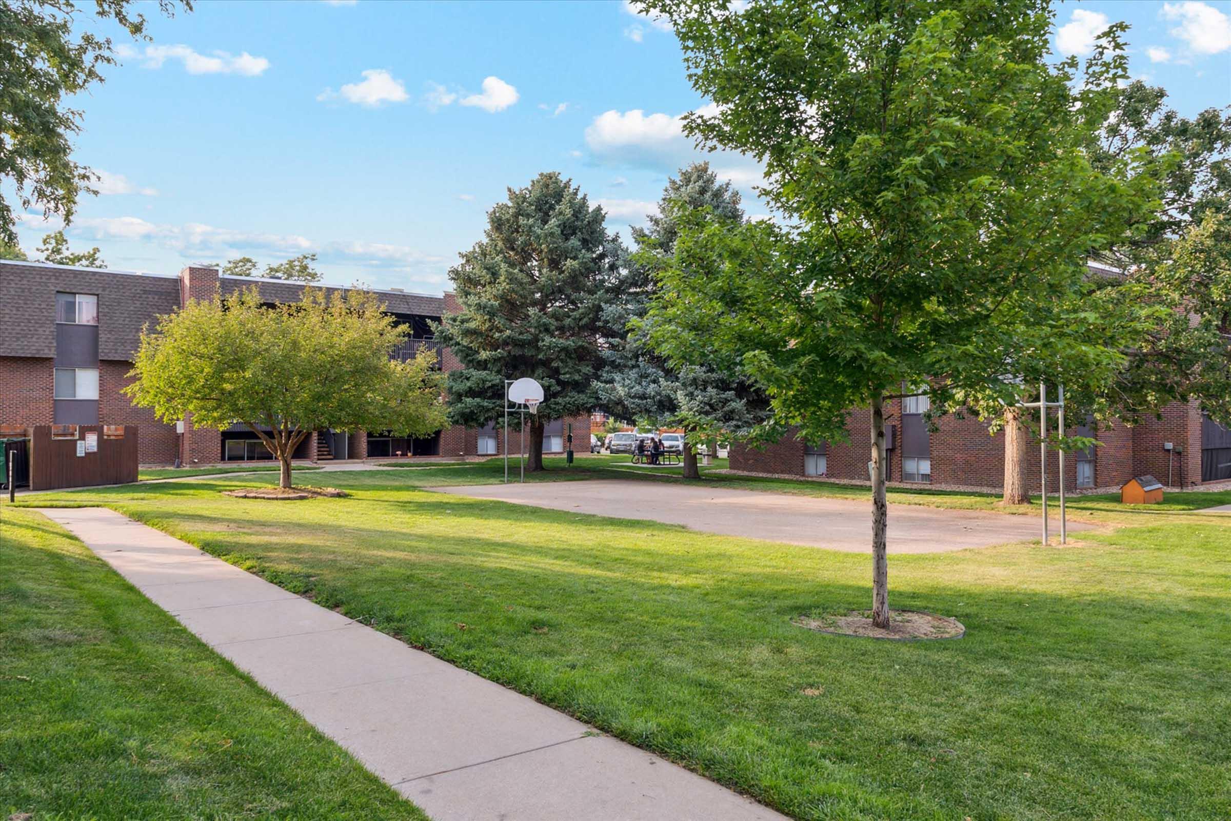 A grassy courtyard featuring a basketball hoop, flanked by trees and two brick apartment buildings. A sidewalk leads through the area, with blue skies and scattered clouds in the background. The setting suggests a peaceful residential neighborhood.