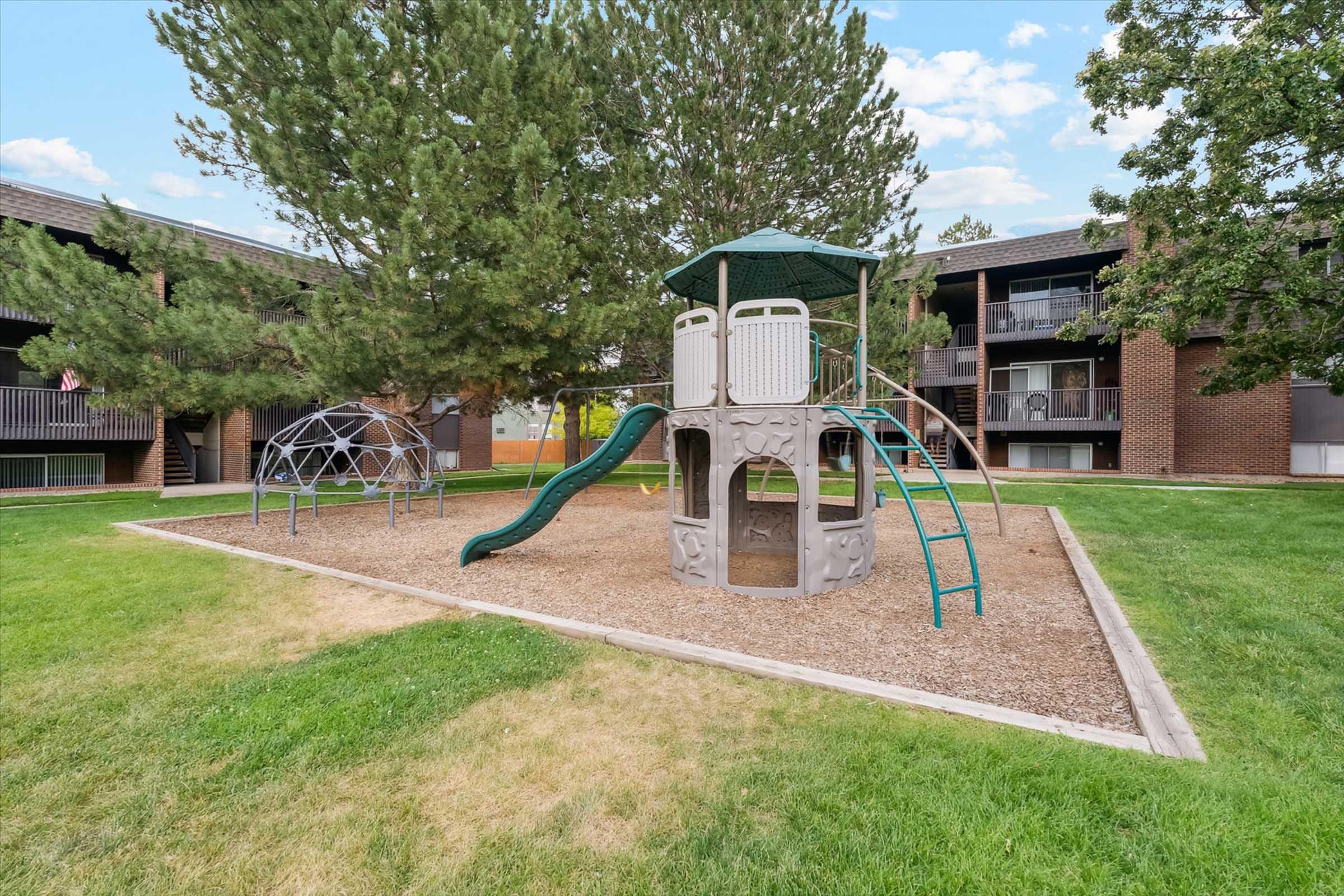 Playground area with a climbing structure featuring slides and a tunnel, surrounded by grassy space. In the background, there are brick apartment buildings and a dome-shaped climbing frame. The scene is set under clear skies with trees providing shade.