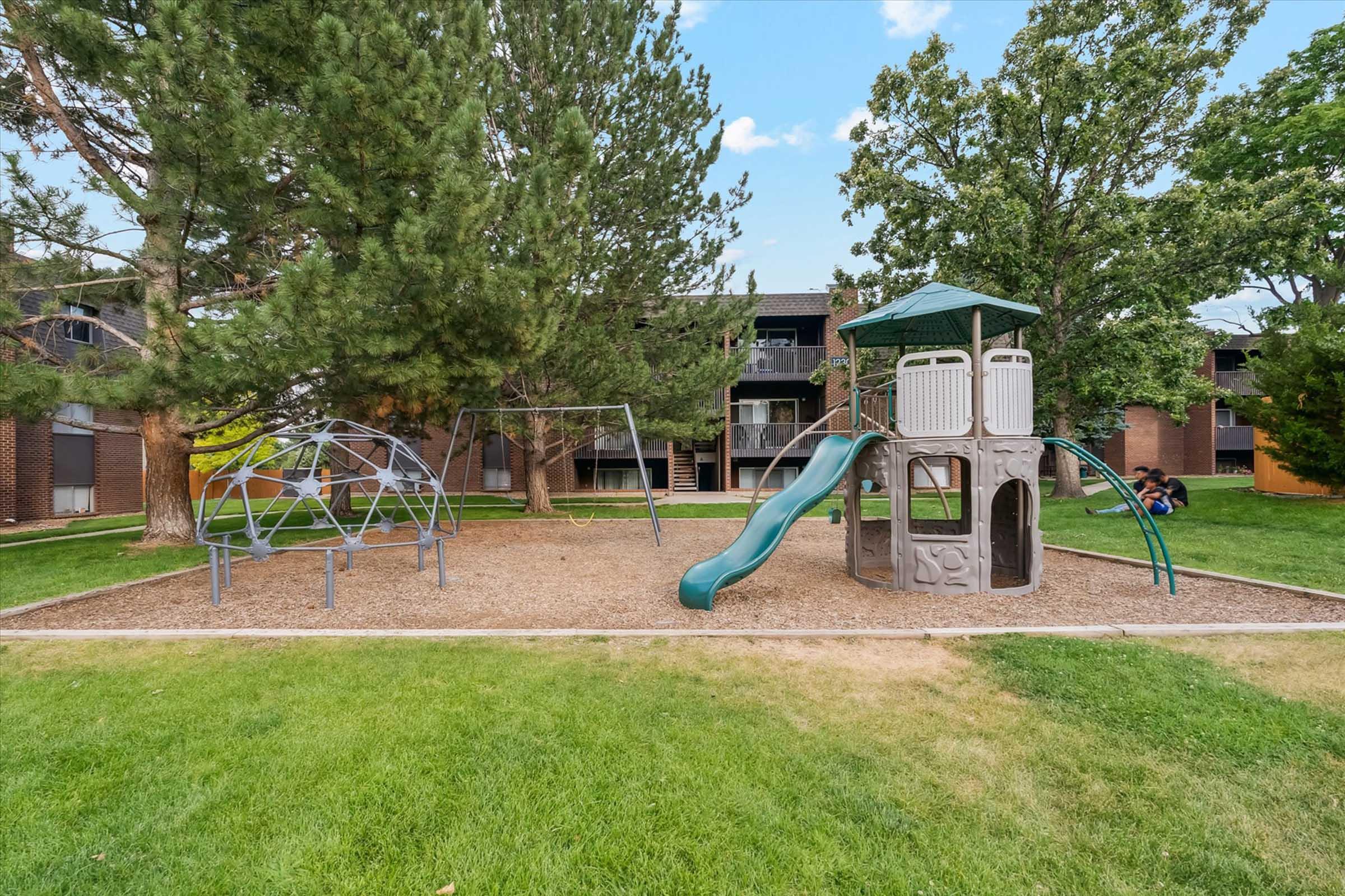 A grassy playground featuring a slide, climbing structure, and swings, surrounded by trees. In the background, there are apartment buildings. The area is sunny and well-maintained, providing a welcoming environment for children to play.