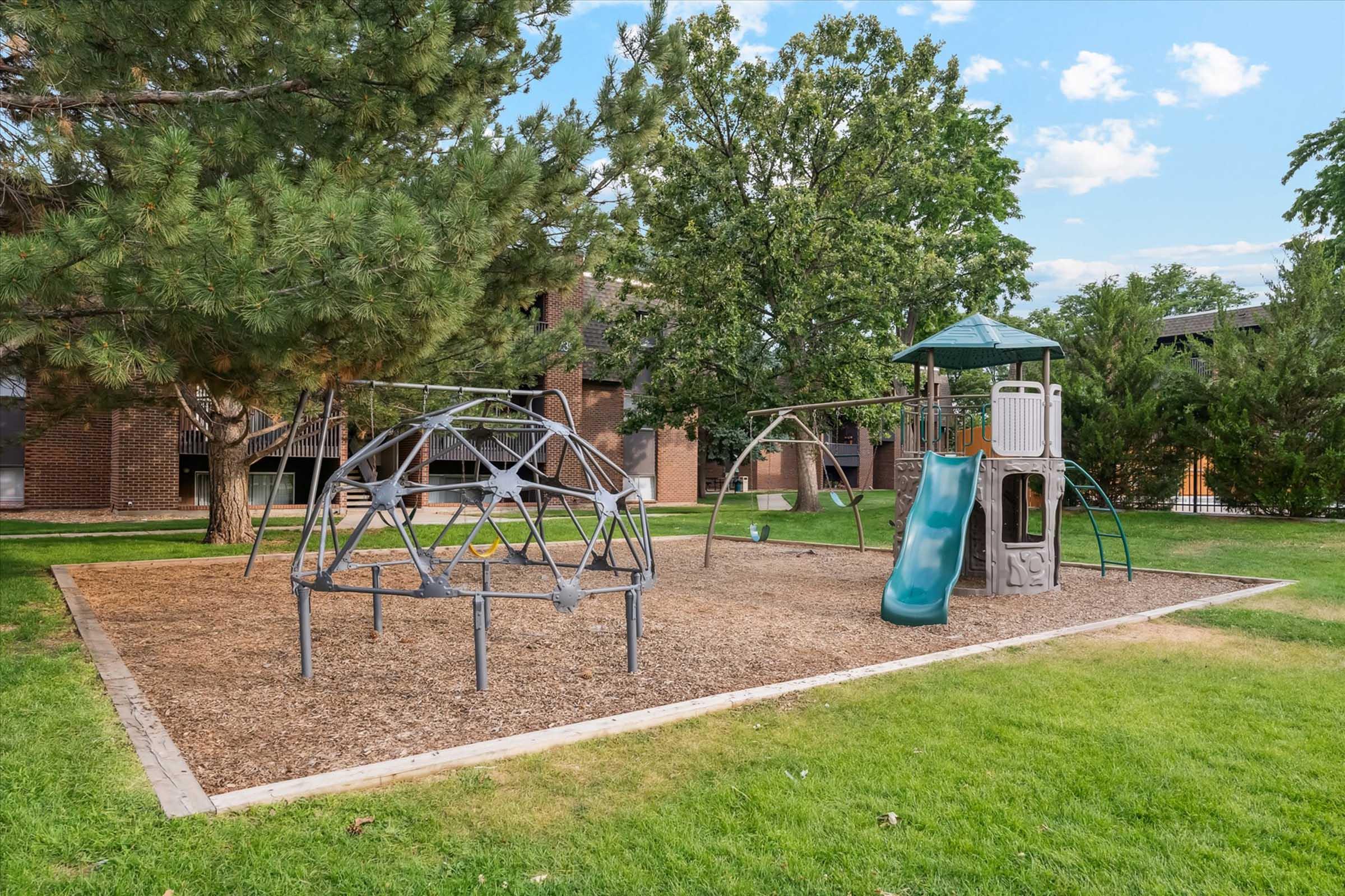Playground area featuring a climbing structure, slide, and swings, surrounded by grass and trees. The ground is covered with wood chips, and apartment buildings are visible in the background under a blue sky with scattered clouds.