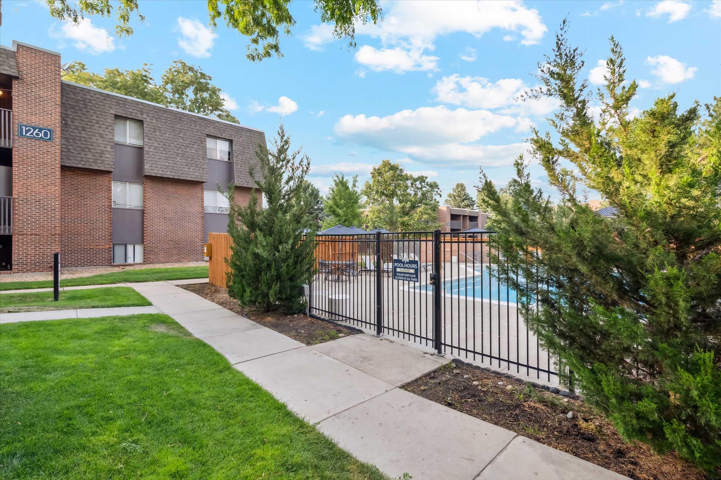A well-maintained outdoor area featuring a fenced swimming pool surrounded by green grass and decorative shrubs. The background includes a three-story brick building with large windows. The scene is bright and inviting, with blue skies and scattered clouds.