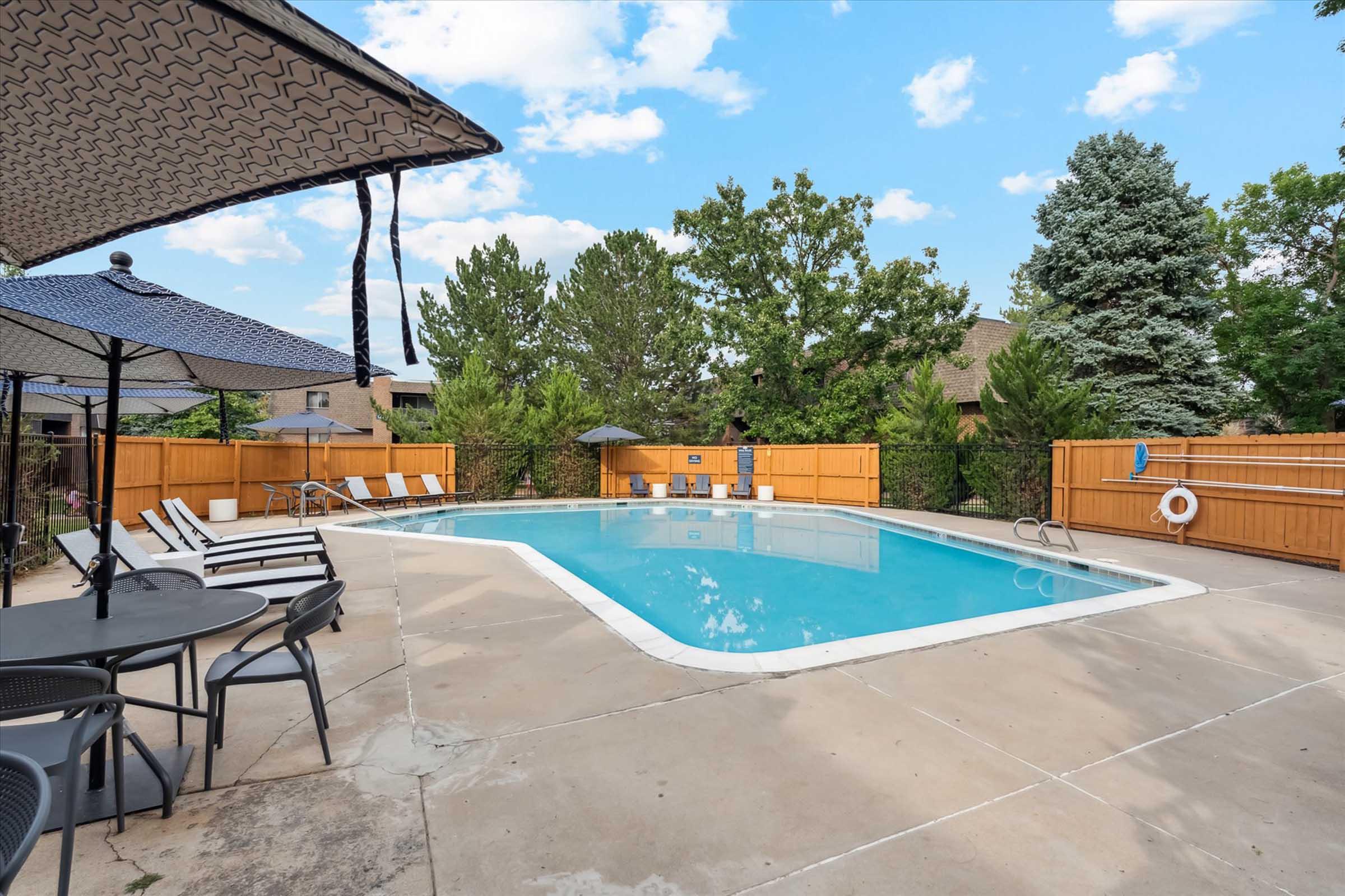 A backyard swimming pool surrounded by a wooden fence. There are lounge chairs and umbrellas set up around the pool, and trees in the background. The sky is partly cloudy, creating a bright and inviting atmosphere.