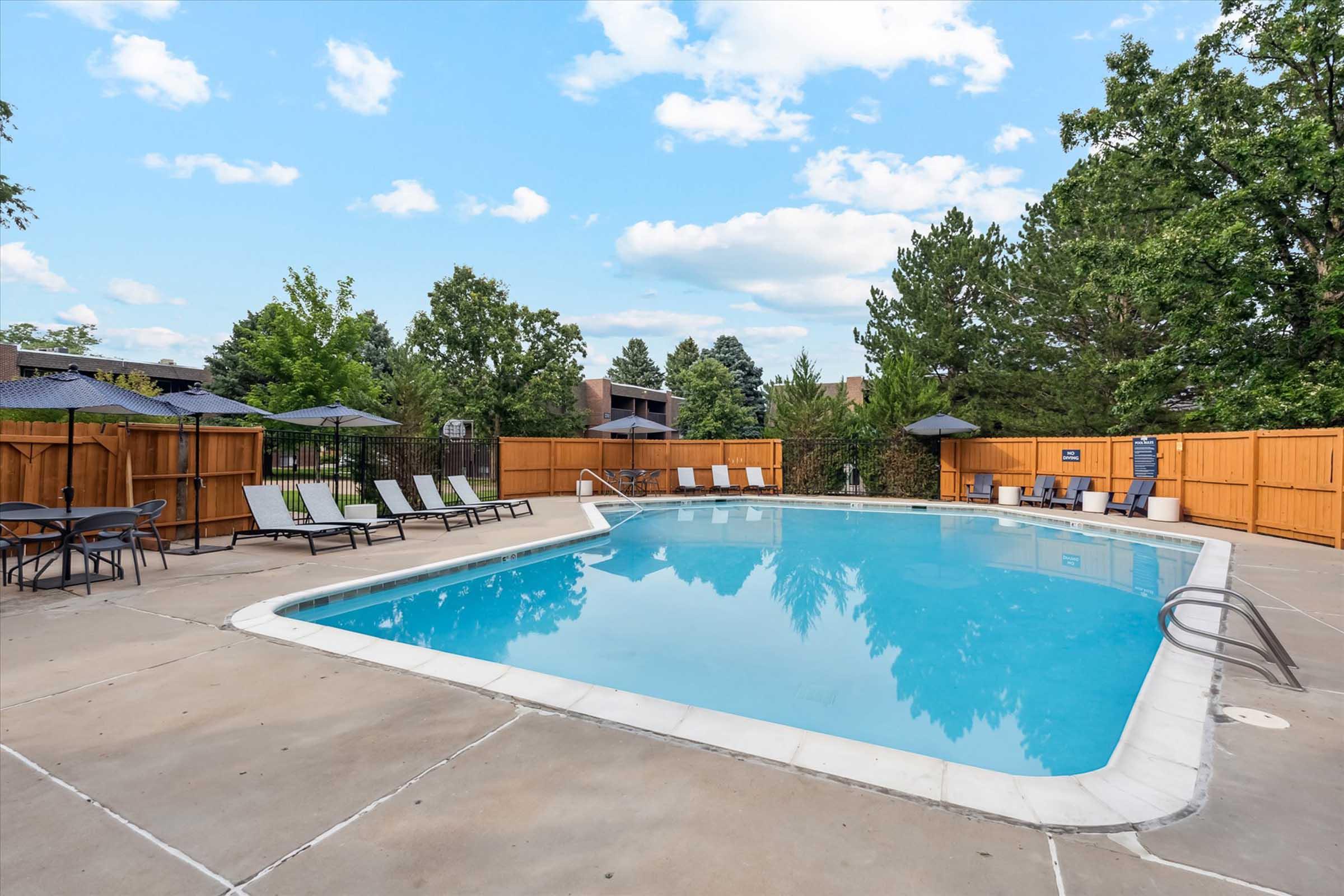 A tranquil outdoor swimming pool surrounded by lounge chairs and shade umbrellas. The pool is bordered by a wooden fence and backed by lush green trees, creating a peaceful atmosphere. The sky is partly cloudy, enhancing the serene setting.