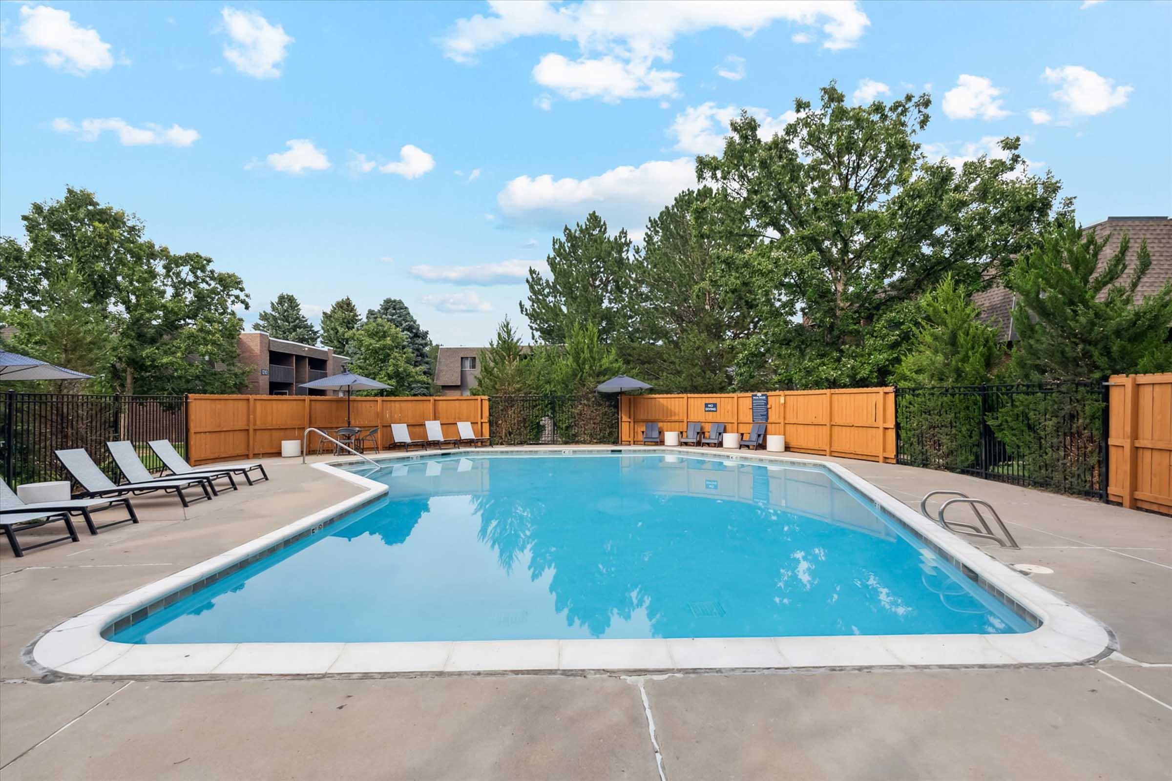 A clean, outdoor swimming pool surrounded by lounge chairs and shaded areas. The pool has clear blue water and is enclosed by a wooden fence, with green trees in the background and a bright blue sky with scattered clouds overhead.