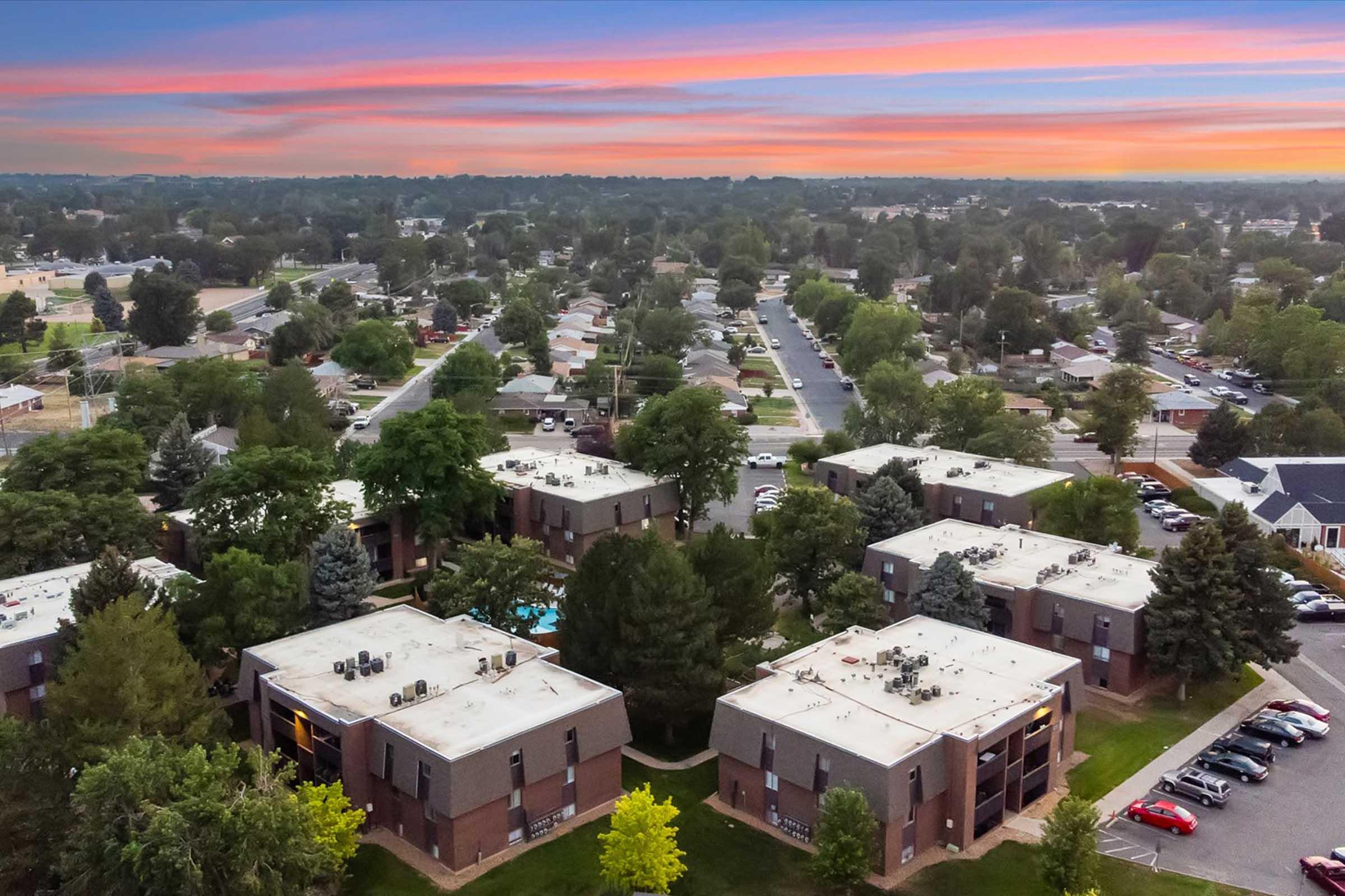 Aerial view of a residential neighborhood featuring multiple low-rise apartment buildings surrounded by trees and greenery. The landscape shows a network of streets and single-family homes, along with a colorful sunset sky in the background.