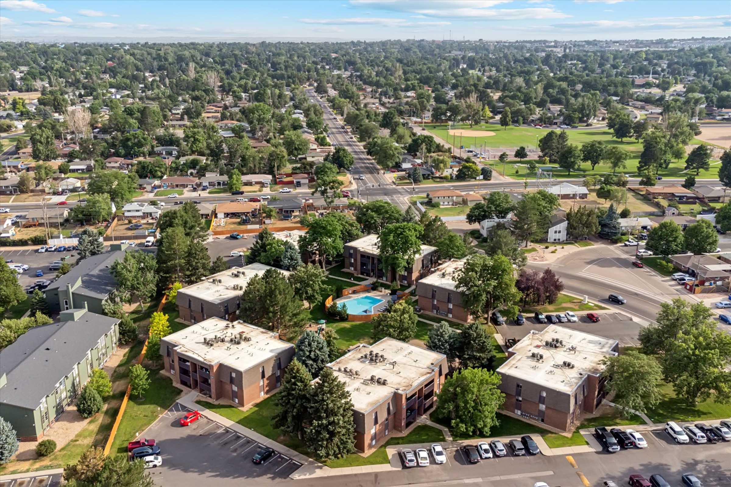 Aerial view of a suburban area featuring several apartment buildings surrounded by trees. A swimming pool is visible in the center of the complex. Nearby, streets are lined with parked cars and green spaces, with a bustling road running through the area. Clear blue sky above.