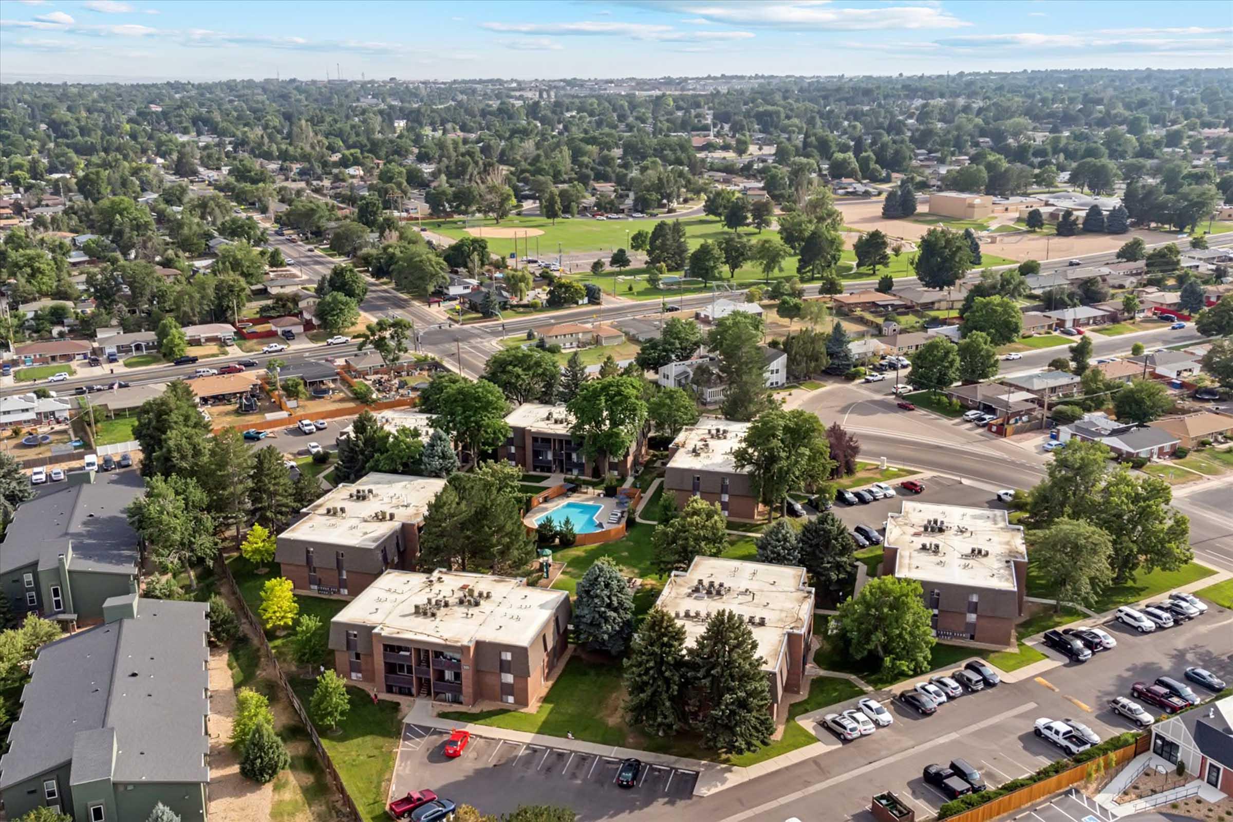 Aerial view of a suburban area featuring multiple apartment buildings with a central pool, surrounded by trees and green parks. In the background, additional residential buildings and streets are visible, along with vehicles parked in the vicinity. The scene showcases a mix of urban and natural elements under a clear blue sky.