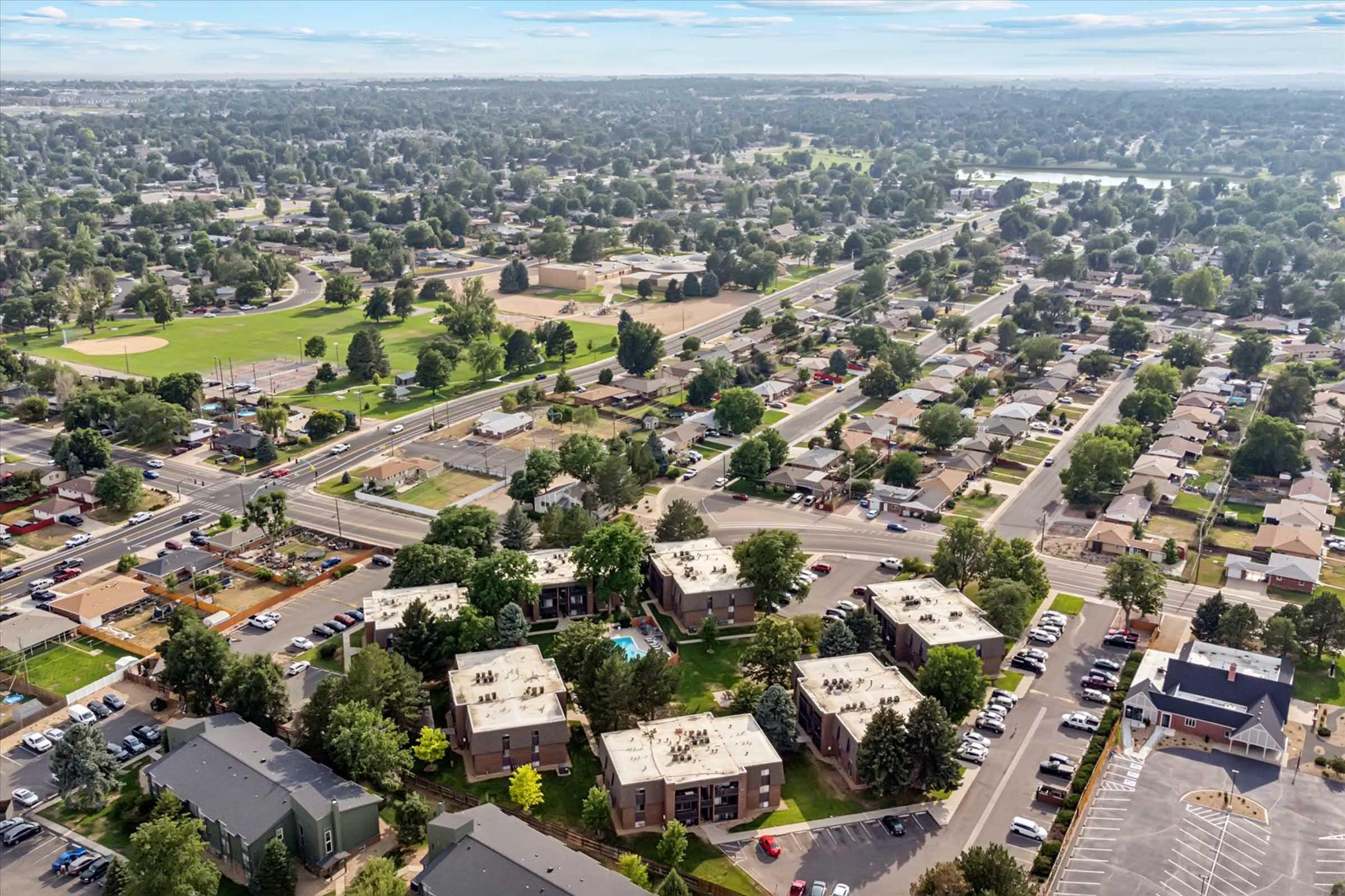 Aerial view of a suburban area featuring residential buildings, parking lots, and green spaces. Roads and pathways are visible, alongside baseball fields and parks. The scene captures a mix of tree-lined streets and neighborhood layouts, showcasing the community's layout and natural surroundings under a clear sky.