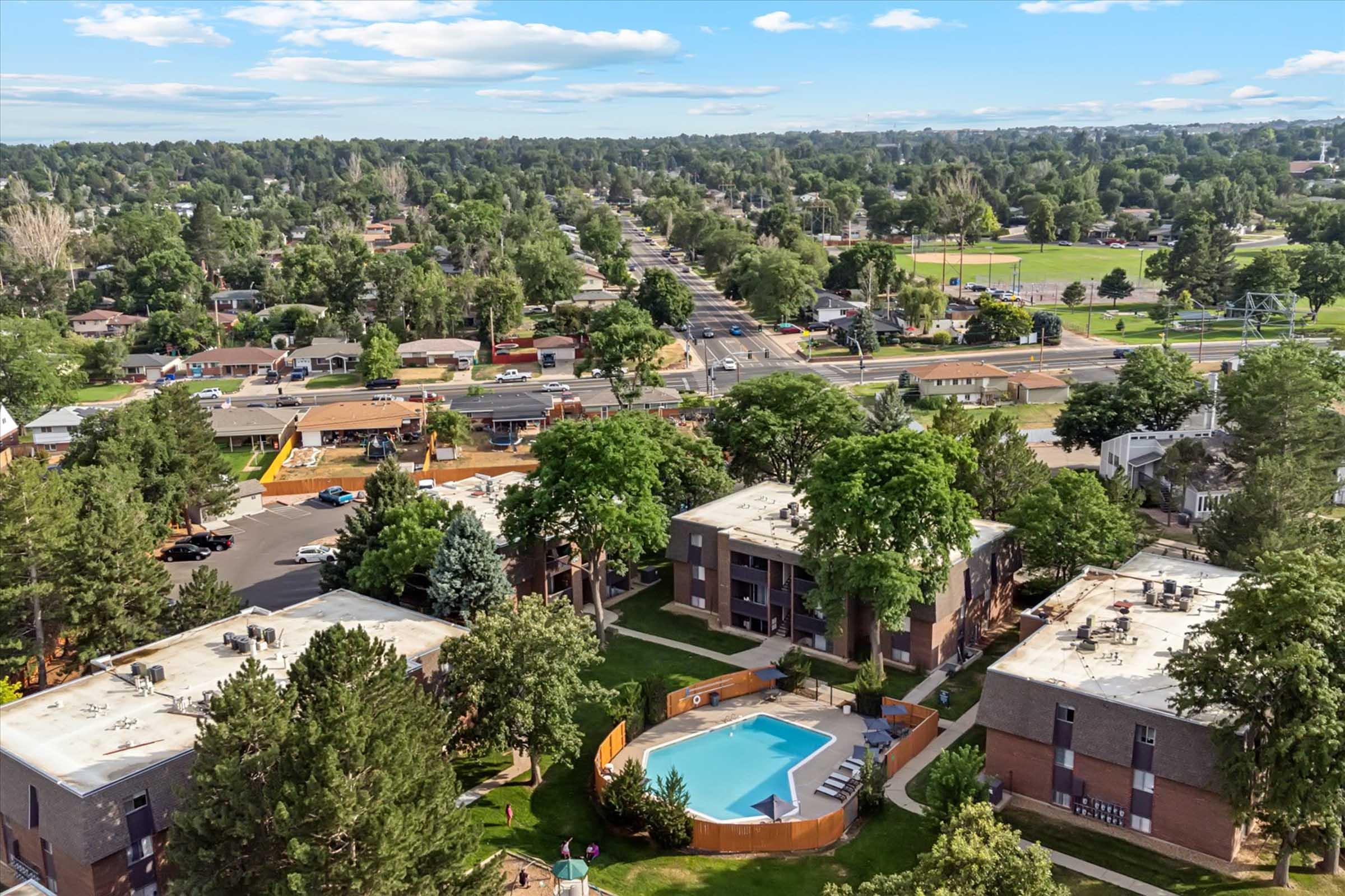 Aerial view of a residential area featuring multiple apartment buildings surrounded by trees. In the foreground, a swimming pool is visible, and the background shows a tree-lined street with houses and green spaces. The sky is partly cloudy, suggesting a pleasant day.