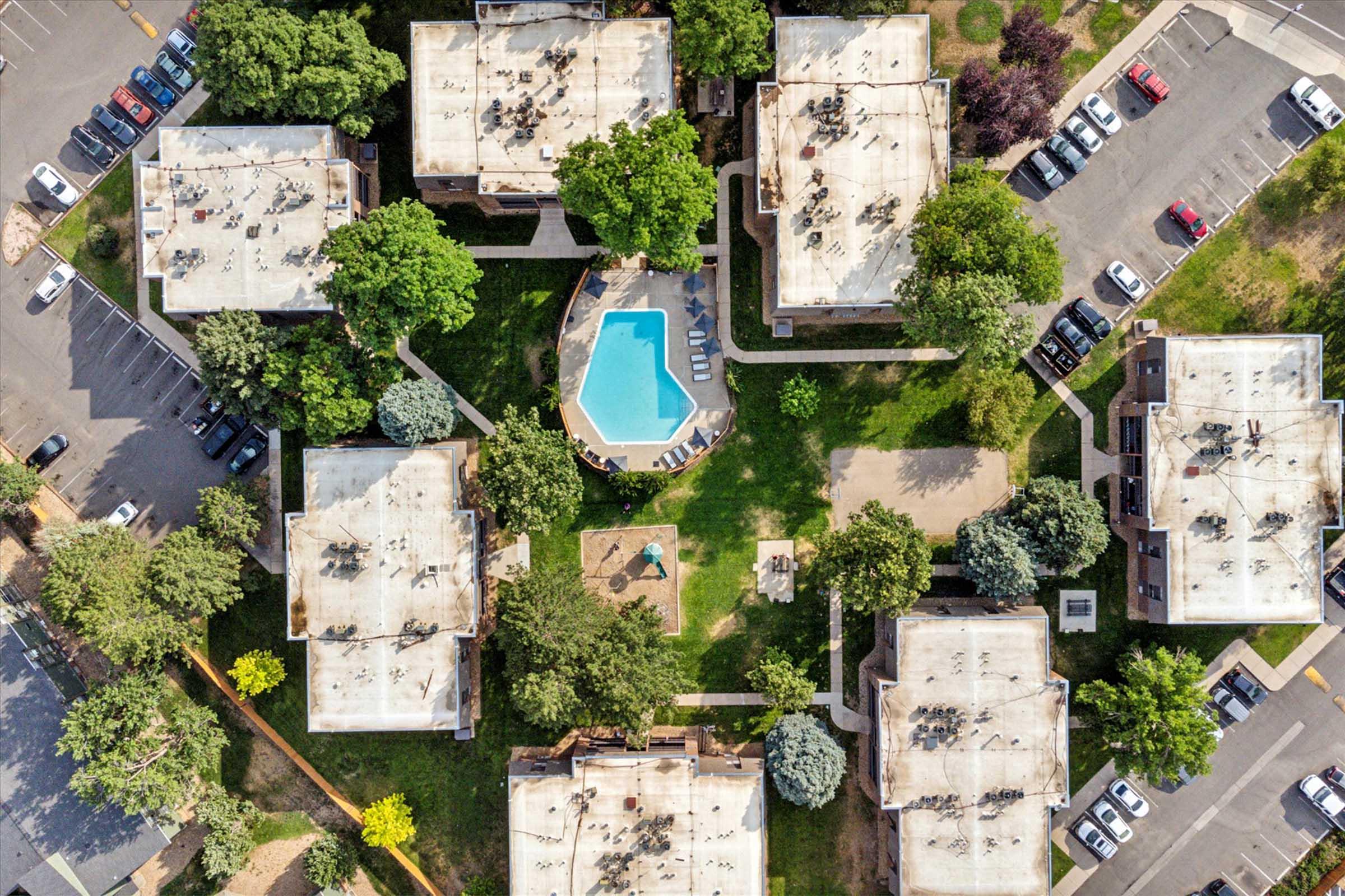 Bird's-eye view of an apartment complex featuring several rectangular buildings arranged around a central pool. The pool area includes lounge chairs and greenery, with pathways connecting the buildings. Surrounding the complex, parking spaces and trees are visible, highlighting a landscaped environment.